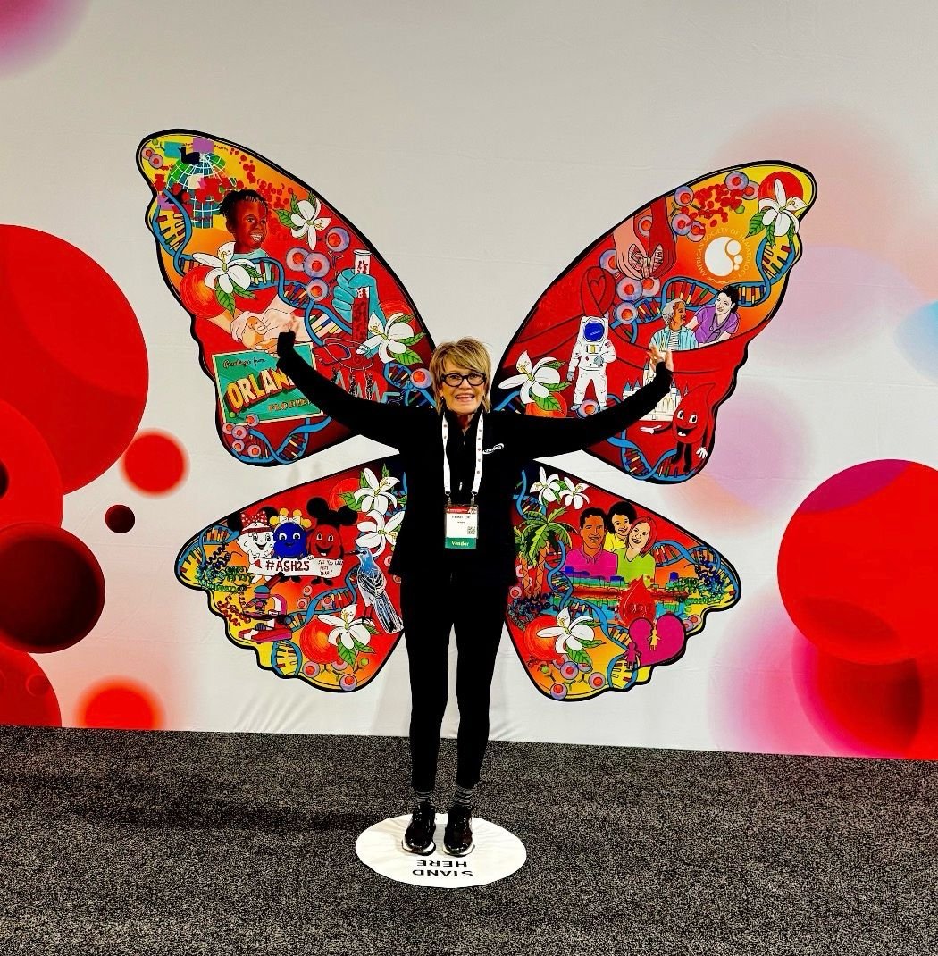 Heather at ASH.A woman with glasses standing with arms raised in front of a large butterfly mural with colorful illustrations of people, animals, and various objects.