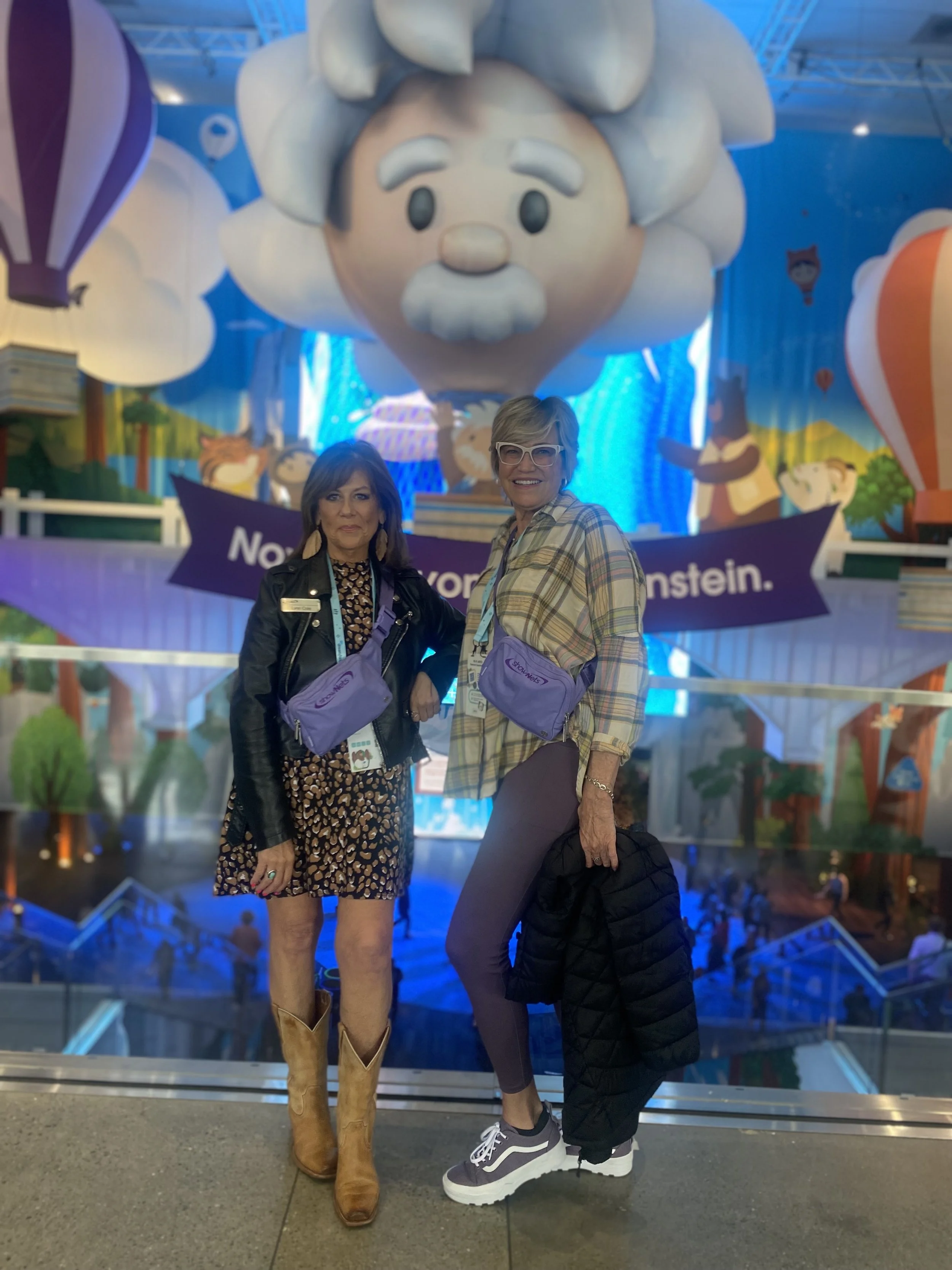 Two women standing together in front of an animated character of Albert Einstein in a colorful, playful setting.