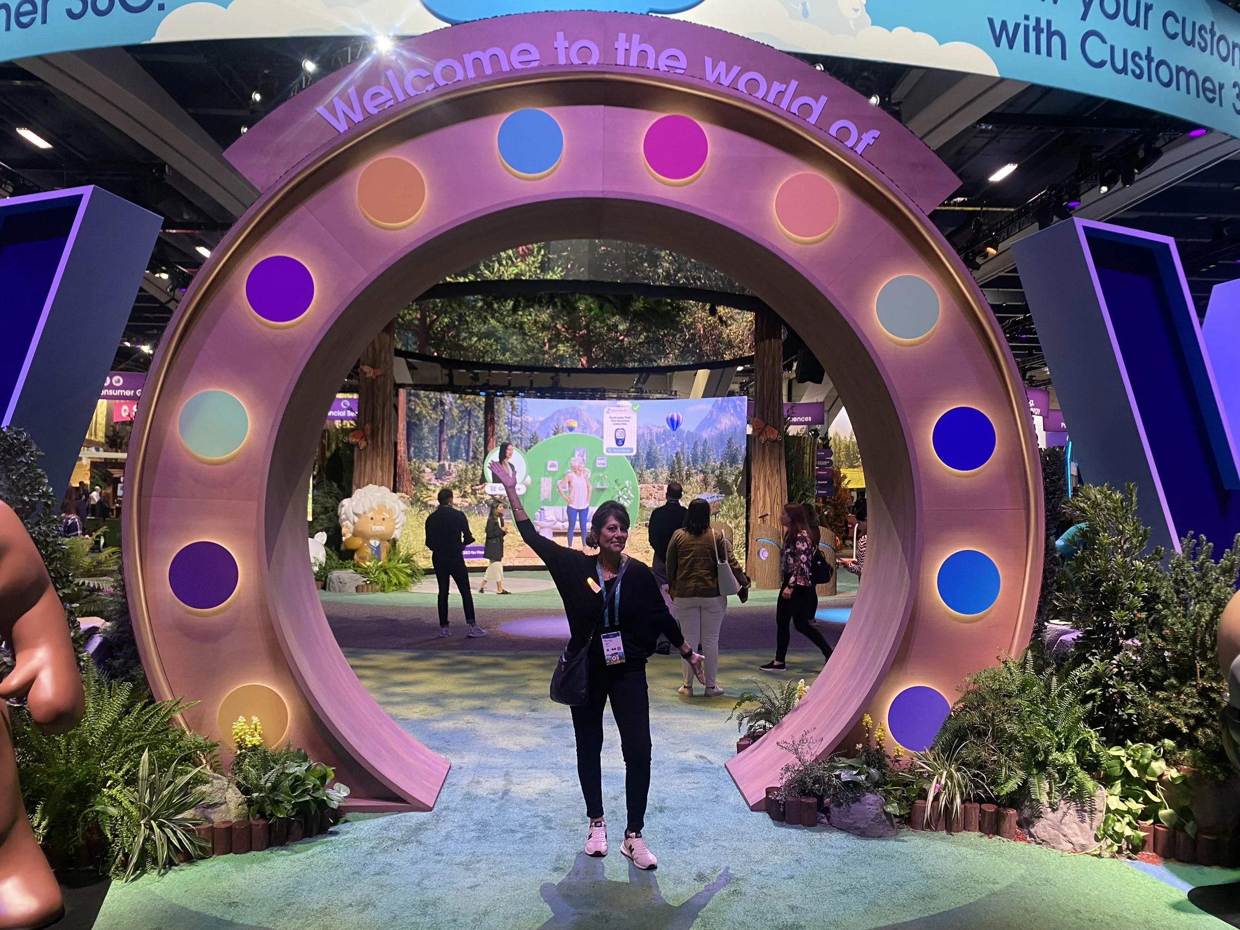 A convention or exhibition with a large circular decorative arch in the foreground, decorated with colorful circles and plants on the sides. In the background, there are people exploring the booths, with a digital screen displaying nature scenery, an