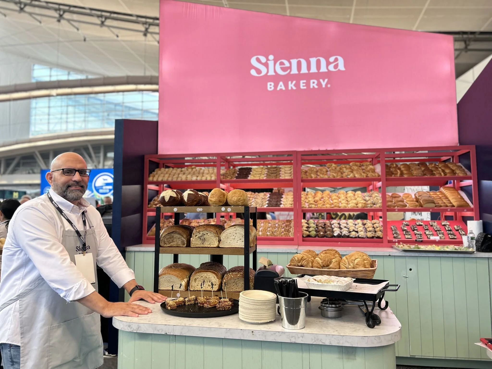 A man wearing glasses and a white shirt stands behind a bakery display counter at an event. The display features various types of bread including sliced bread, rolls, and baguettes. Behind him are shelves filled with assorted baked goods, and a large