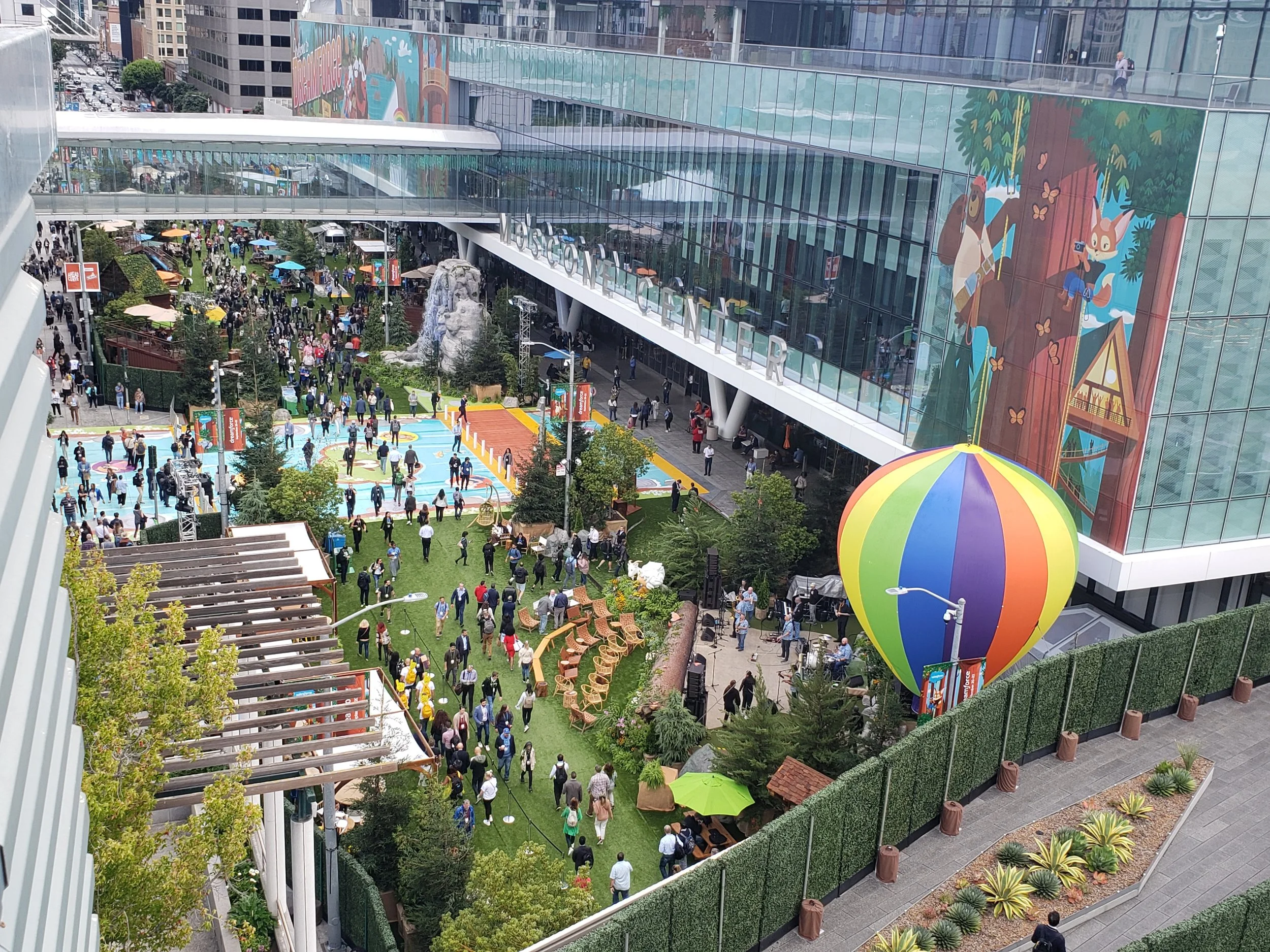 Crowd gathered in outdoor event space at a modern building with large glass windows and colorful mural art, featuring a playground and seating areas with umbrellas.