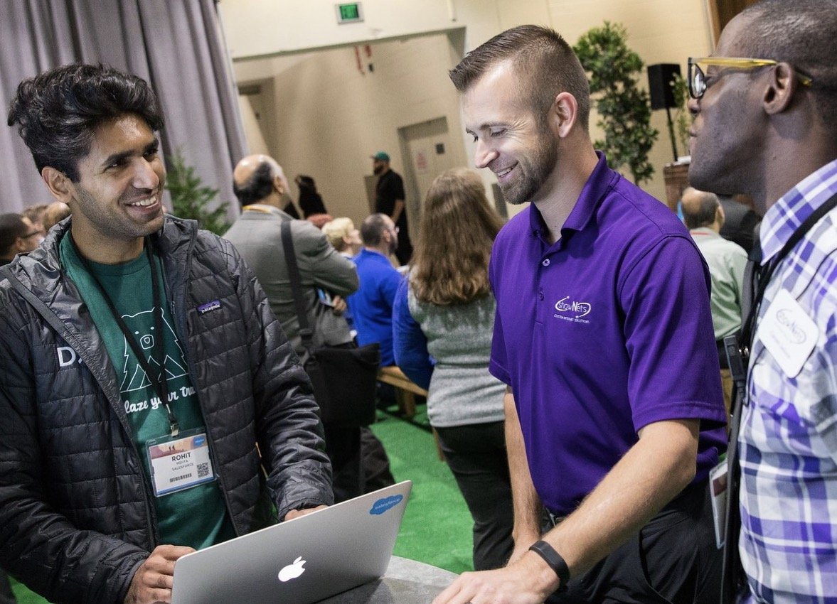 Three men are engaged in conversation at a professional event. The man on the left wears a black jacket and has a name tag that reads "Rohit." The man in the middle is in a purple polo shirt with a logo, and the man on the right is in a checkered shirt with glasses. They are standing in front of a laptop, smiling, with other people and event setting behind them.