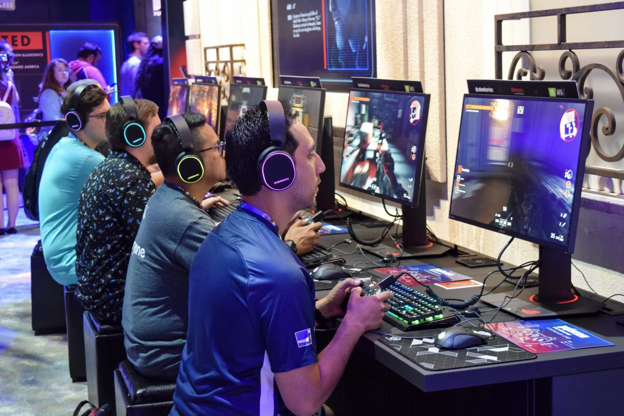 People playing video games at a gaming event, wearing headsets and using controllers on a long table with multiple computer screens.