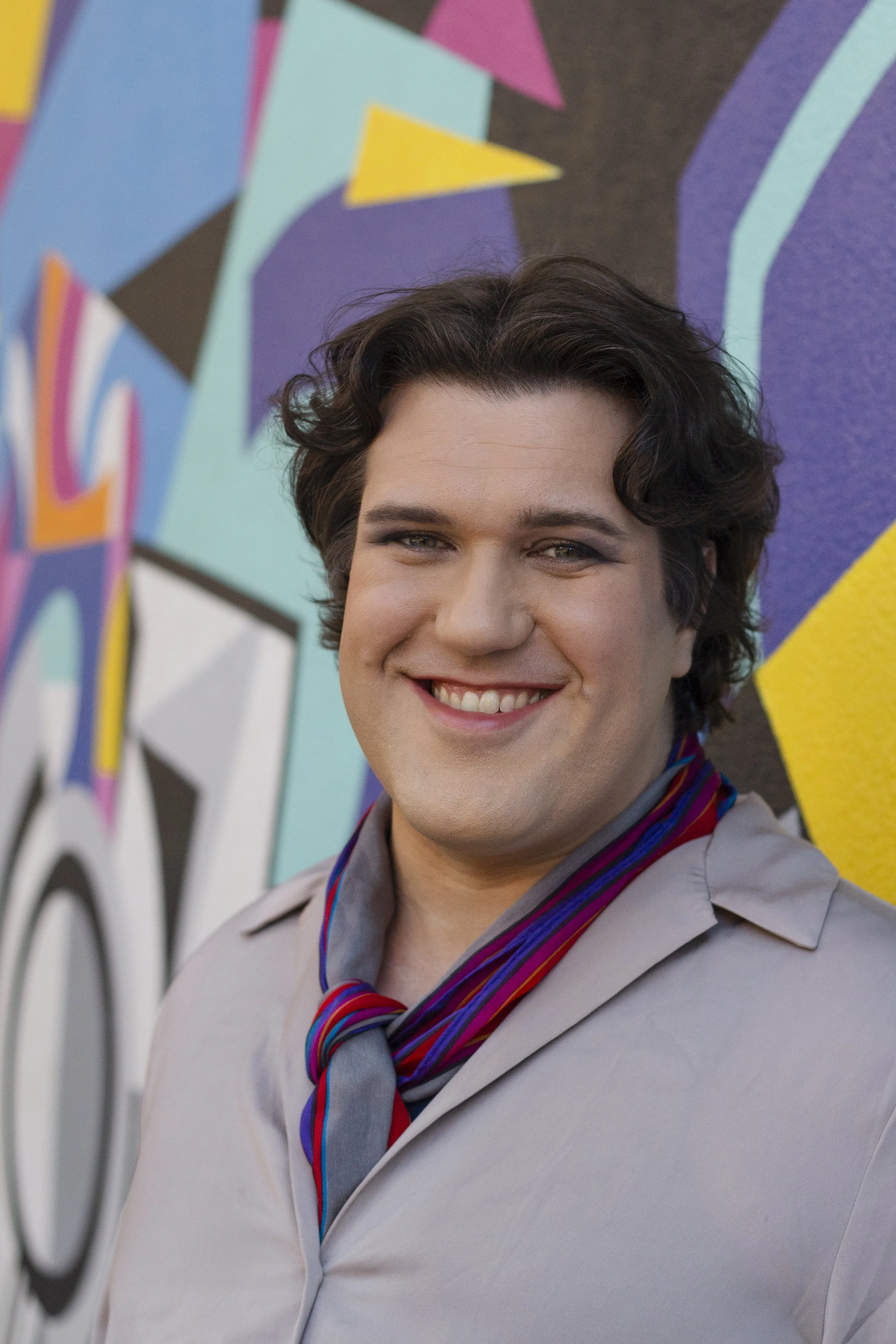 A person smiling in front of a colorful abstract mural with geometric shapes.