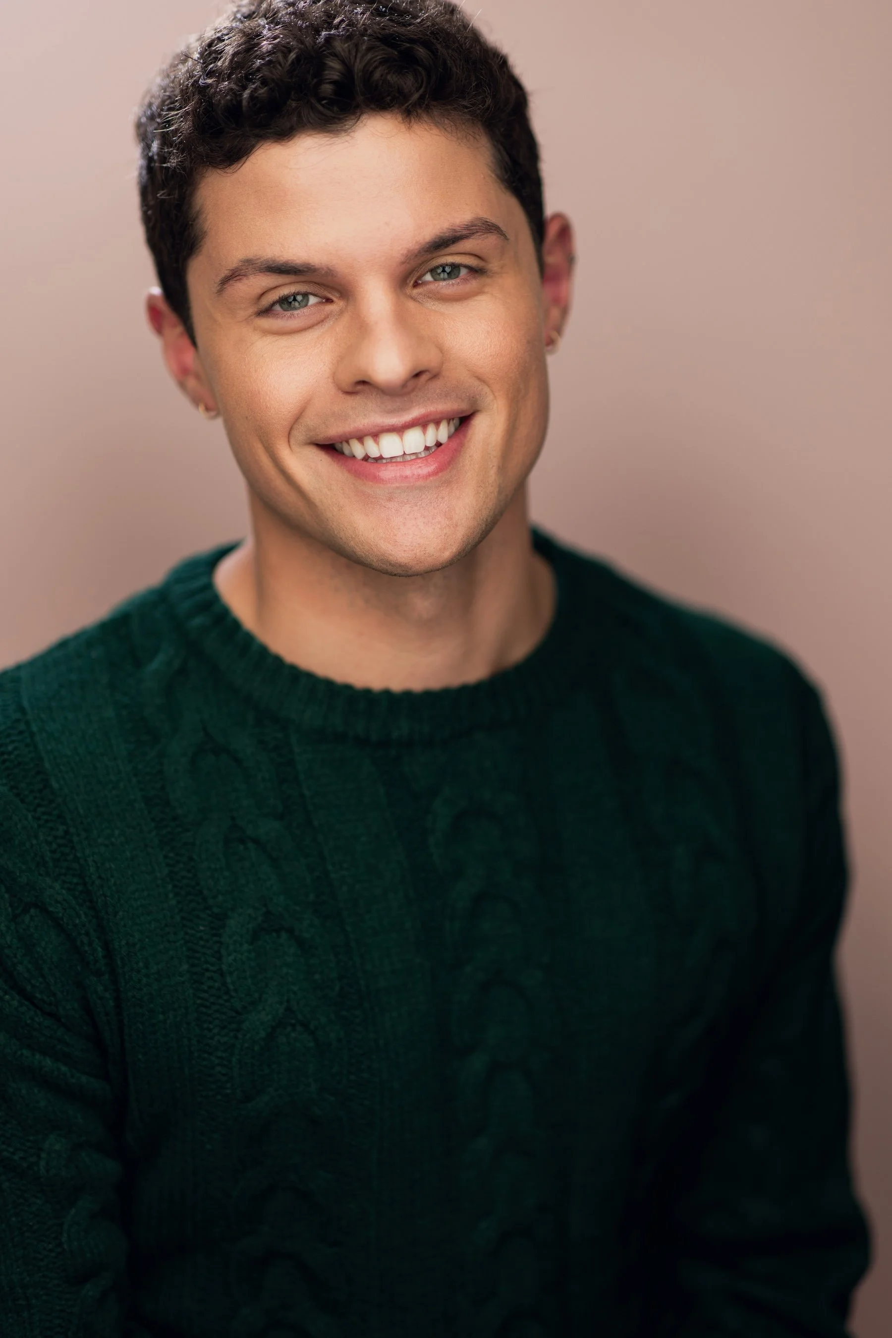 Close-up portrait of a young man with dark curly hair, light blue eyes, and a bright smile, wearing a dark green knitted sweater against a neutral background.