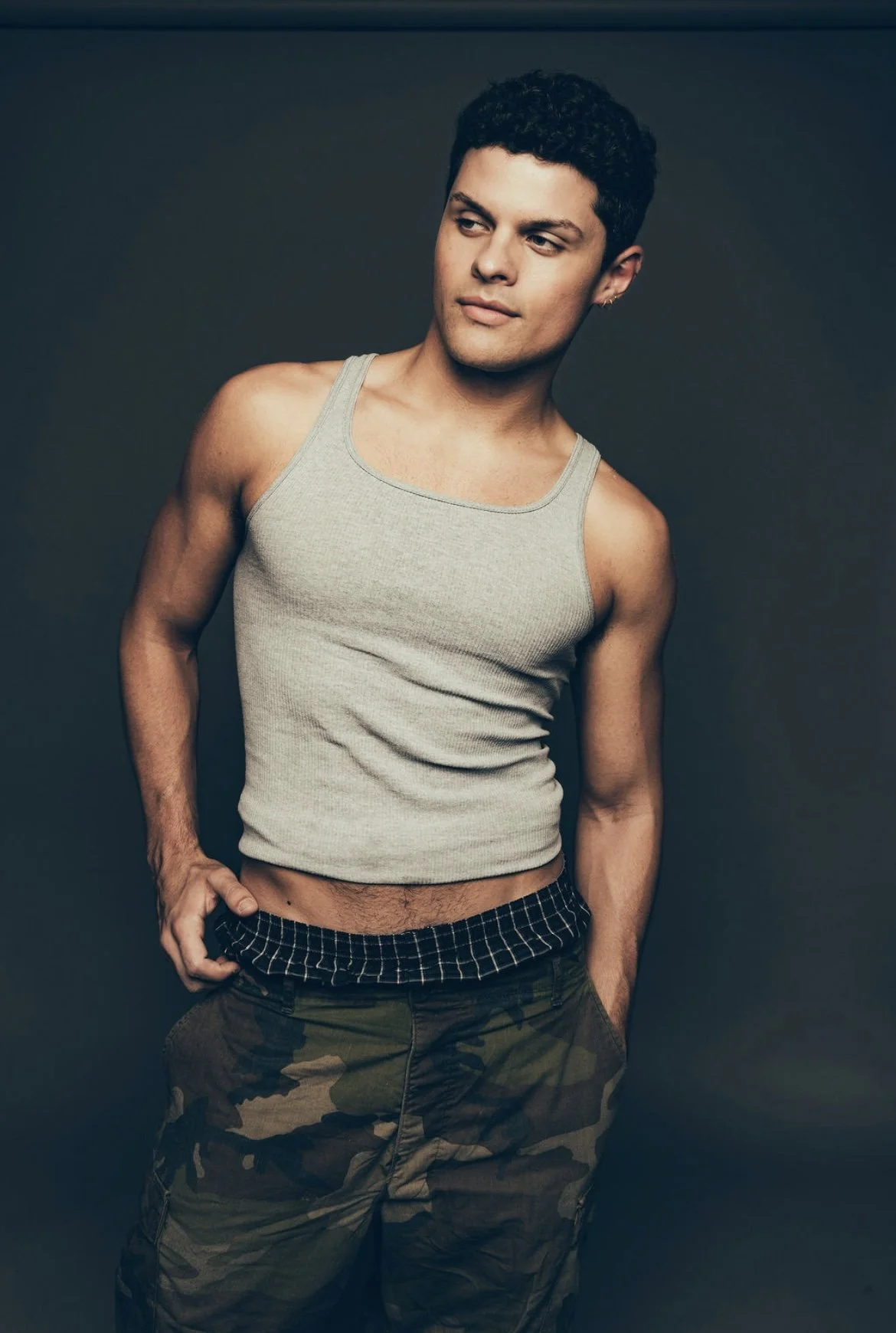 A young man with dark curly hair and light skin, wearing a gray tank top and camouflage pants, standing against a dark background. He has one hand in his pocket and is looking to the side with a slight smile.