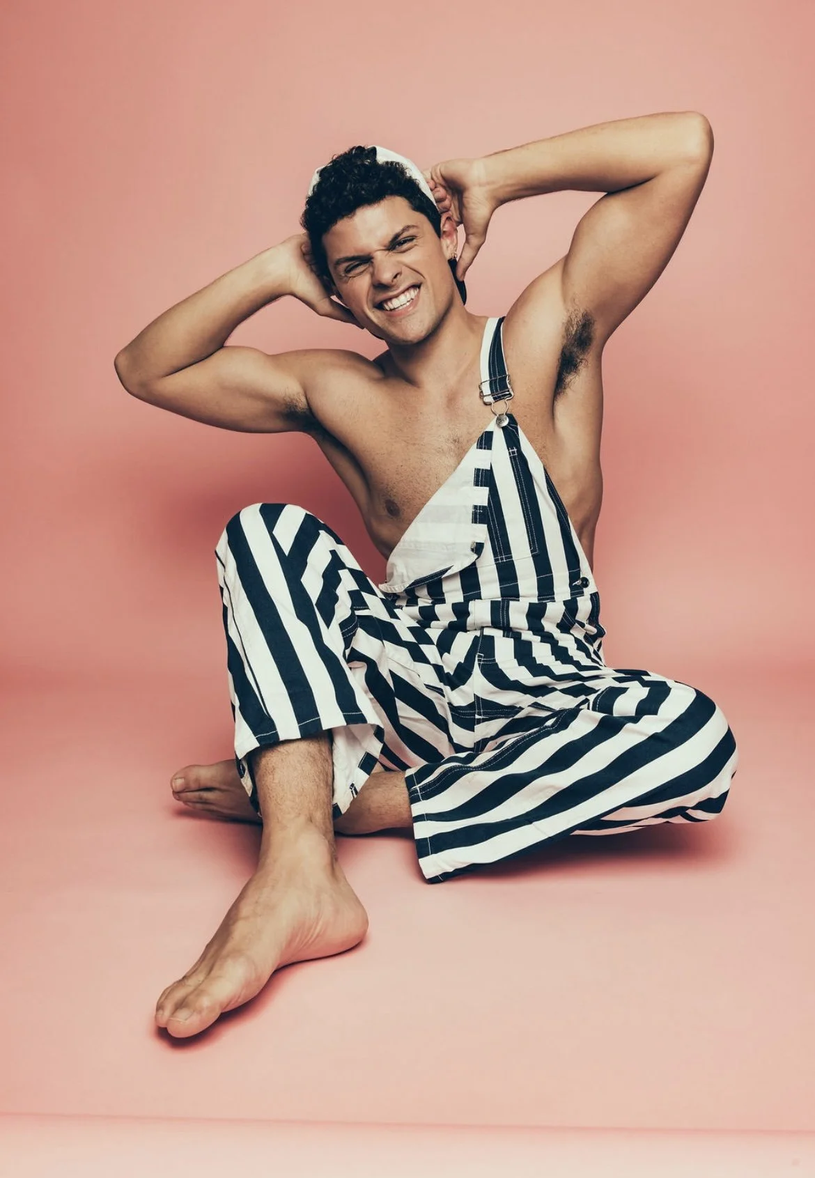 Young man with curly hair, sitting on a pink floor against a pink background, wearing striped overalls, smiling, with arms raised behind his head.