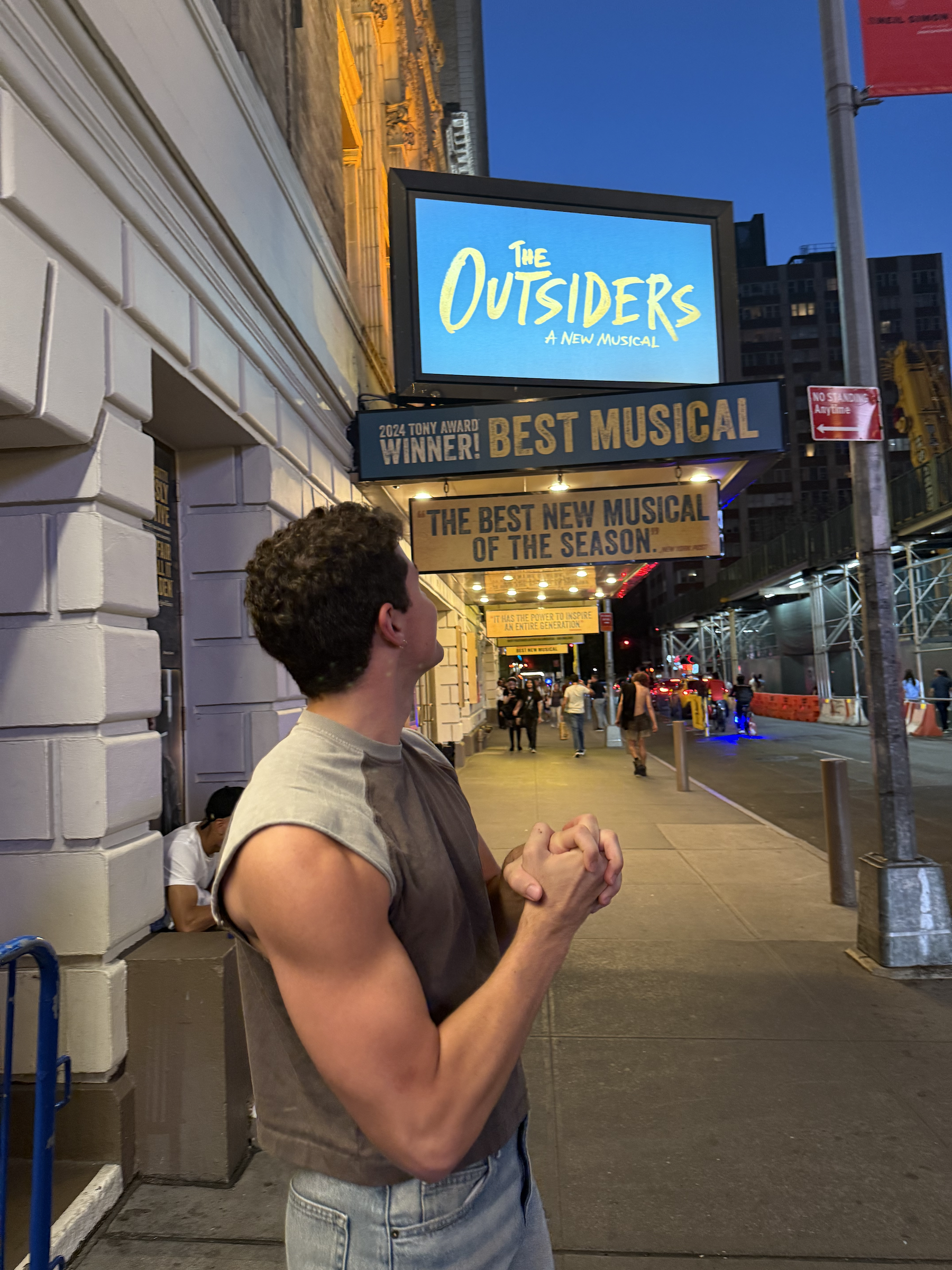 A man with curly hair wearing a gray sleeveless shirt and jeans is standing on a city sidewalk at dusk. He is facing away from the camera, with his hands clasped in front of him. Behind him, there are a few other people and illuminated signs, includi