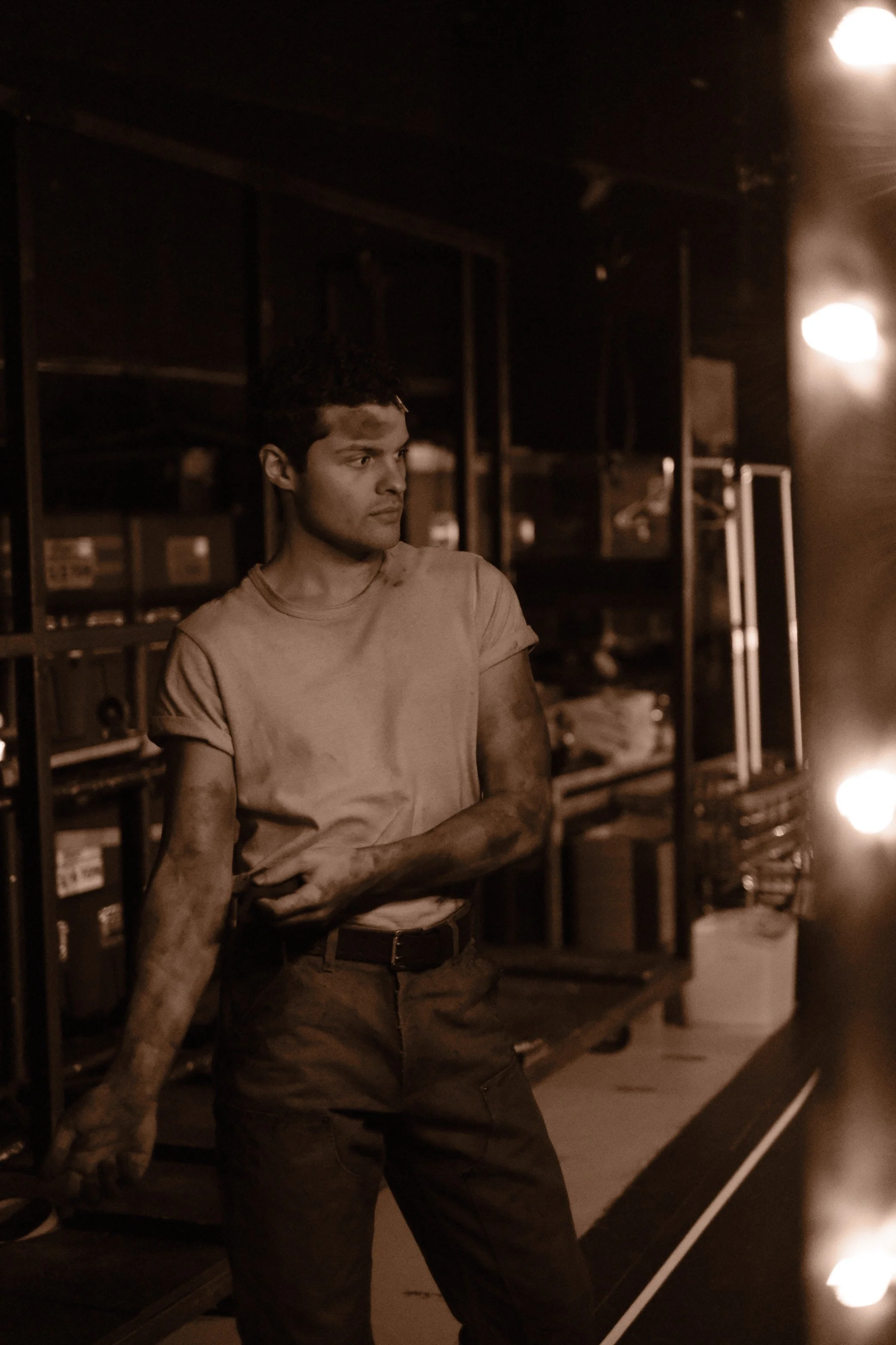 A young man standing in a dimly lit room with industrial shelving in the background, wearing a dirty t-shirt and dark pants.