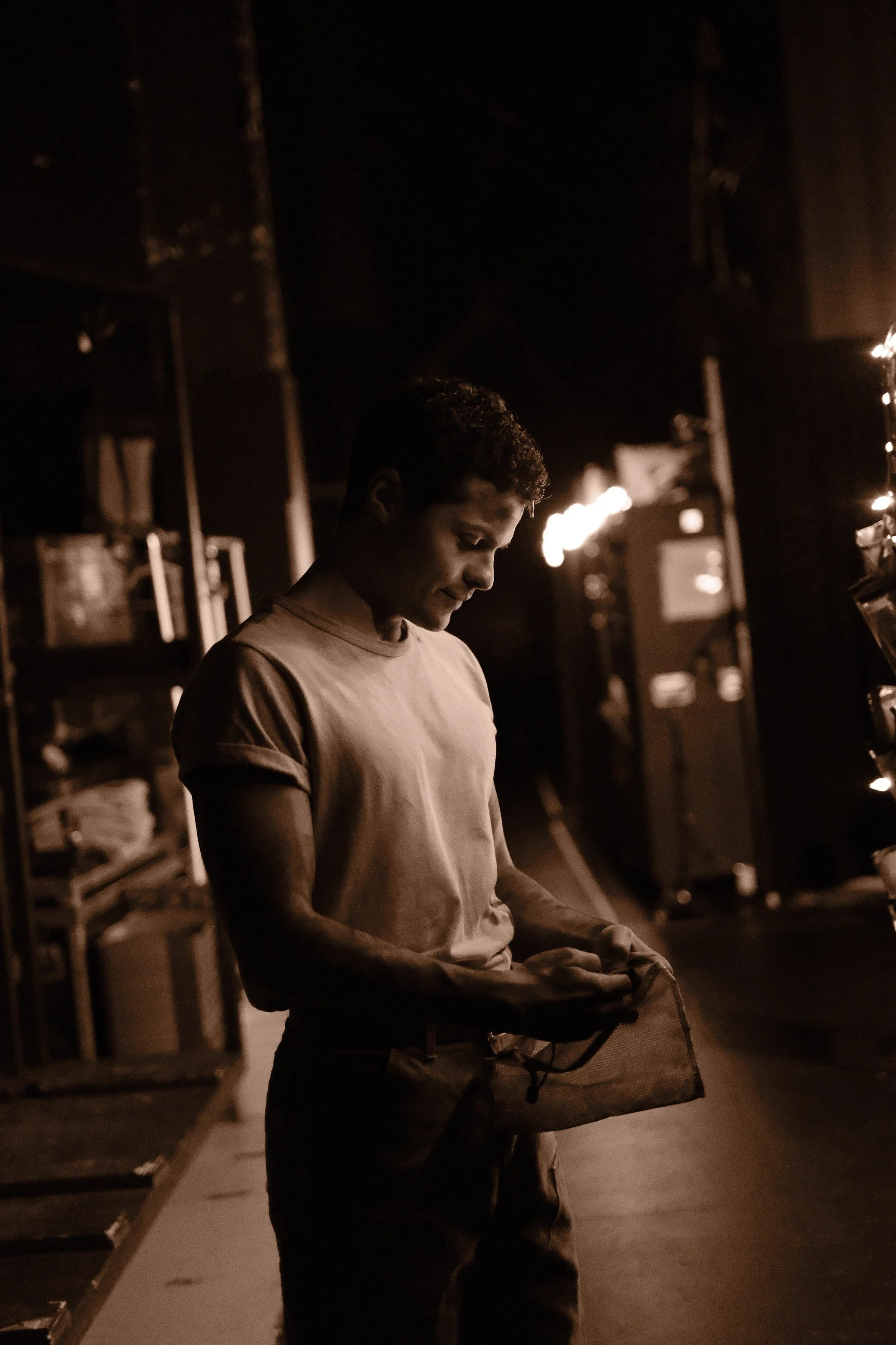 A young man standing in a dimly lit room, looking down at a paper or object in his hands, with shelves and equipment in the background, illuminated by warm light.