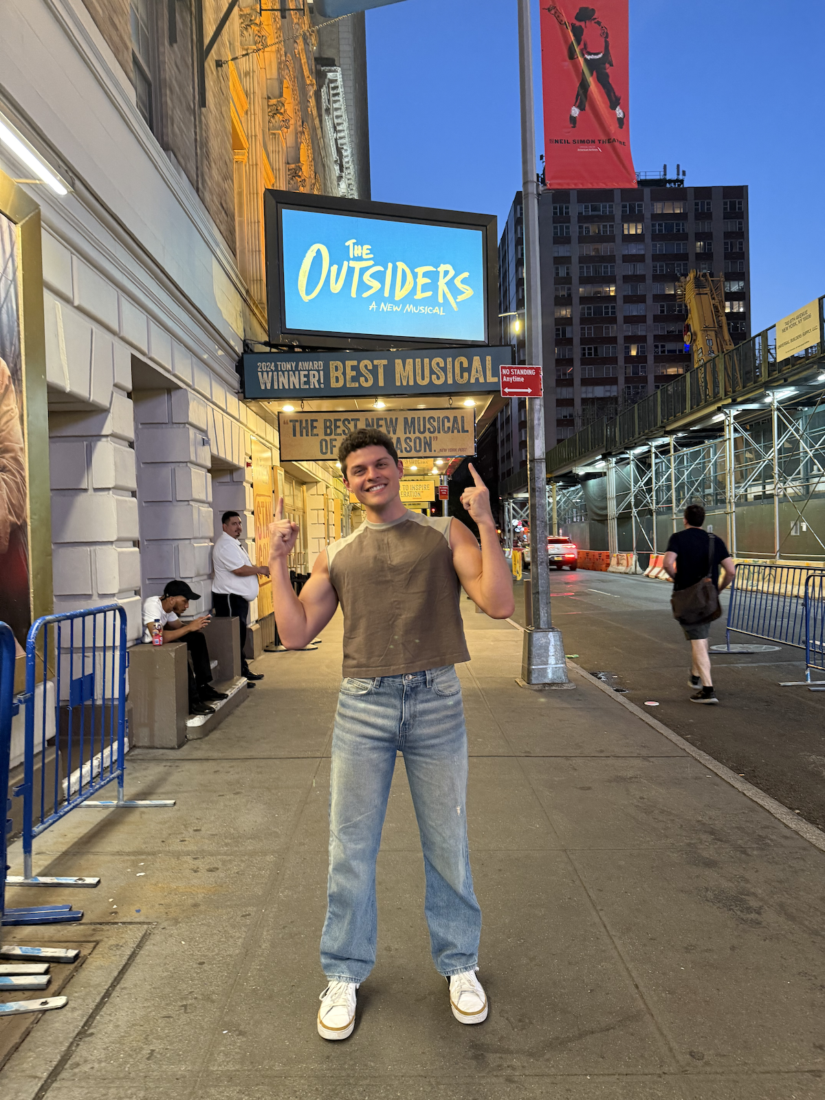 A young man with short dark hair wearing a sleeveless brown top, blue jeans, and white sneakers, standing on a city sidewalk at dusk. He is smiling and pointing upward with both hands. Behind him is a large illuminated sign for the musical 'The Outsi