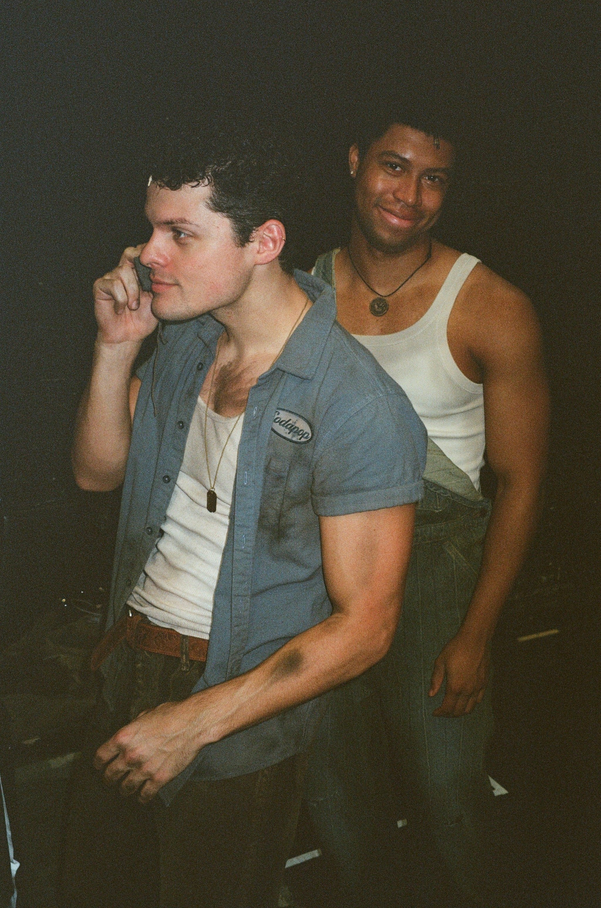 Two men sitting closely together against a dark background. The man on the left has curly dark hair, light skin, and is holding a phone to his ear, wearing a gray shirt with a name patch. The man on the right has short hair, darker skin, and is smili