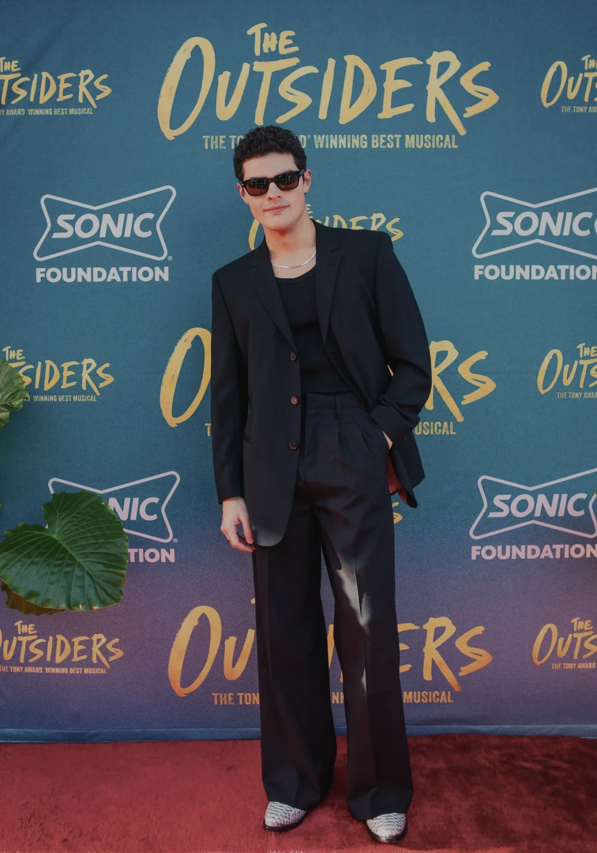 Person wearing sunglasses and a black suit standing on a red carpet in front of a backdrop for the musical 'The Outsiders' with the Sonic Foundation logo.