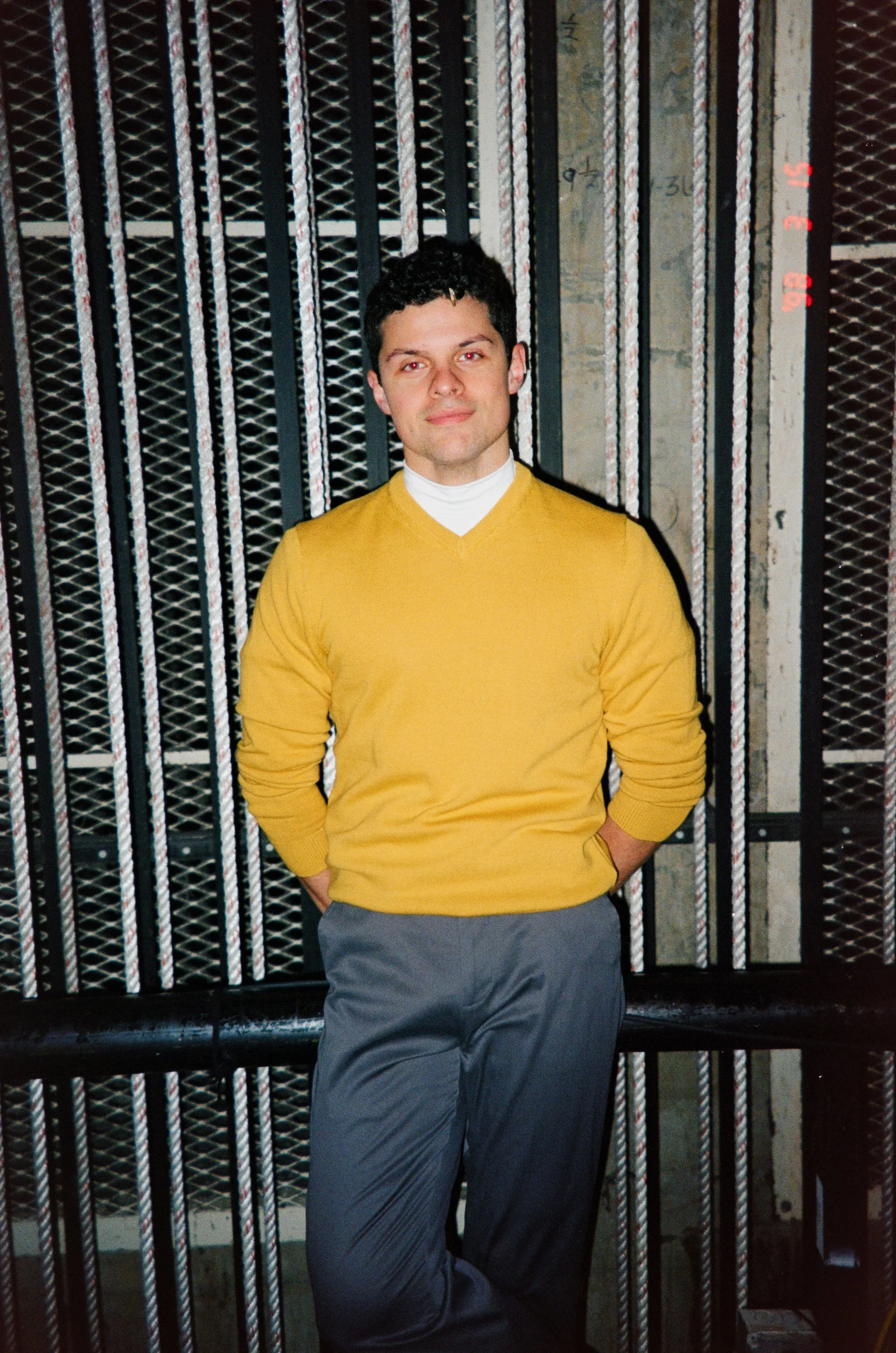 A man wearing a yellow sweater and gray pants standing against a metal garage or elevator gate.