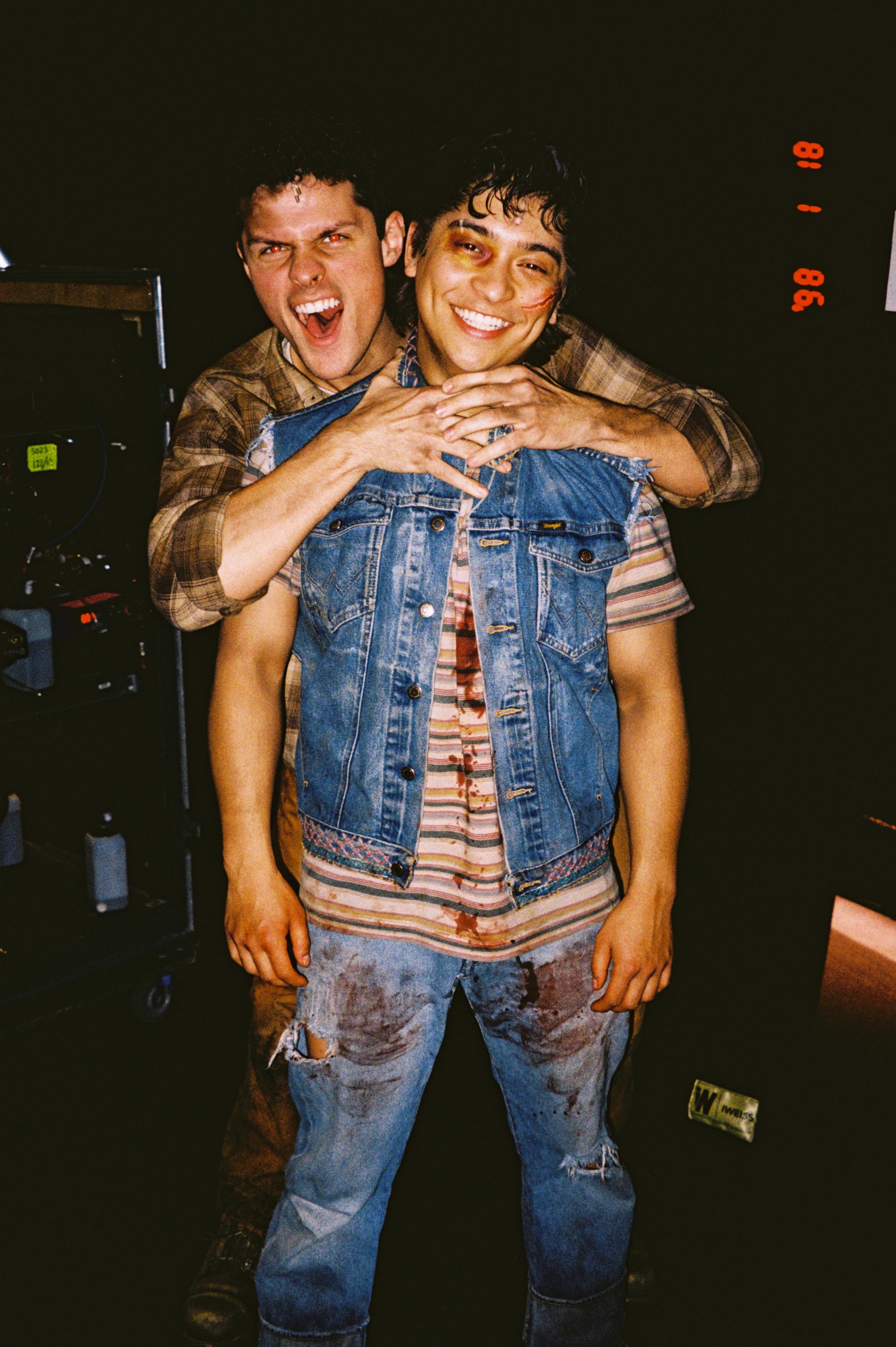Two men in distressed and casual clothing smiling and posing together in a dark room, with one man behind the other. One man has a black eye and blood on his shirt. There is a camera and recording equipment on the left side of the image.