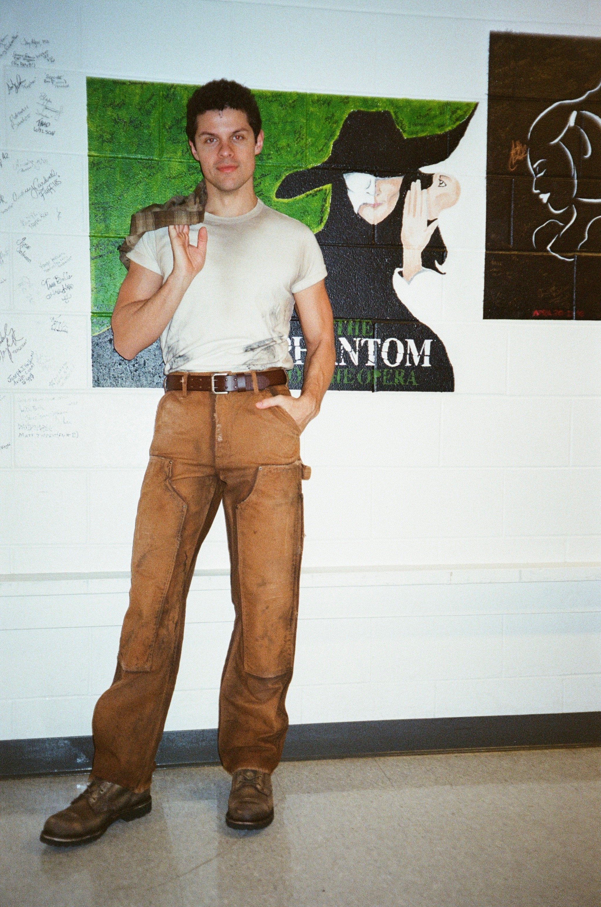 A young man with dark curly hair standing in front of a wall with artwork. He is wearing a light-colored T-shirt, brown pants, and brown boots, holding a brown jacket over his shoulder, with his left hand in his pocket.