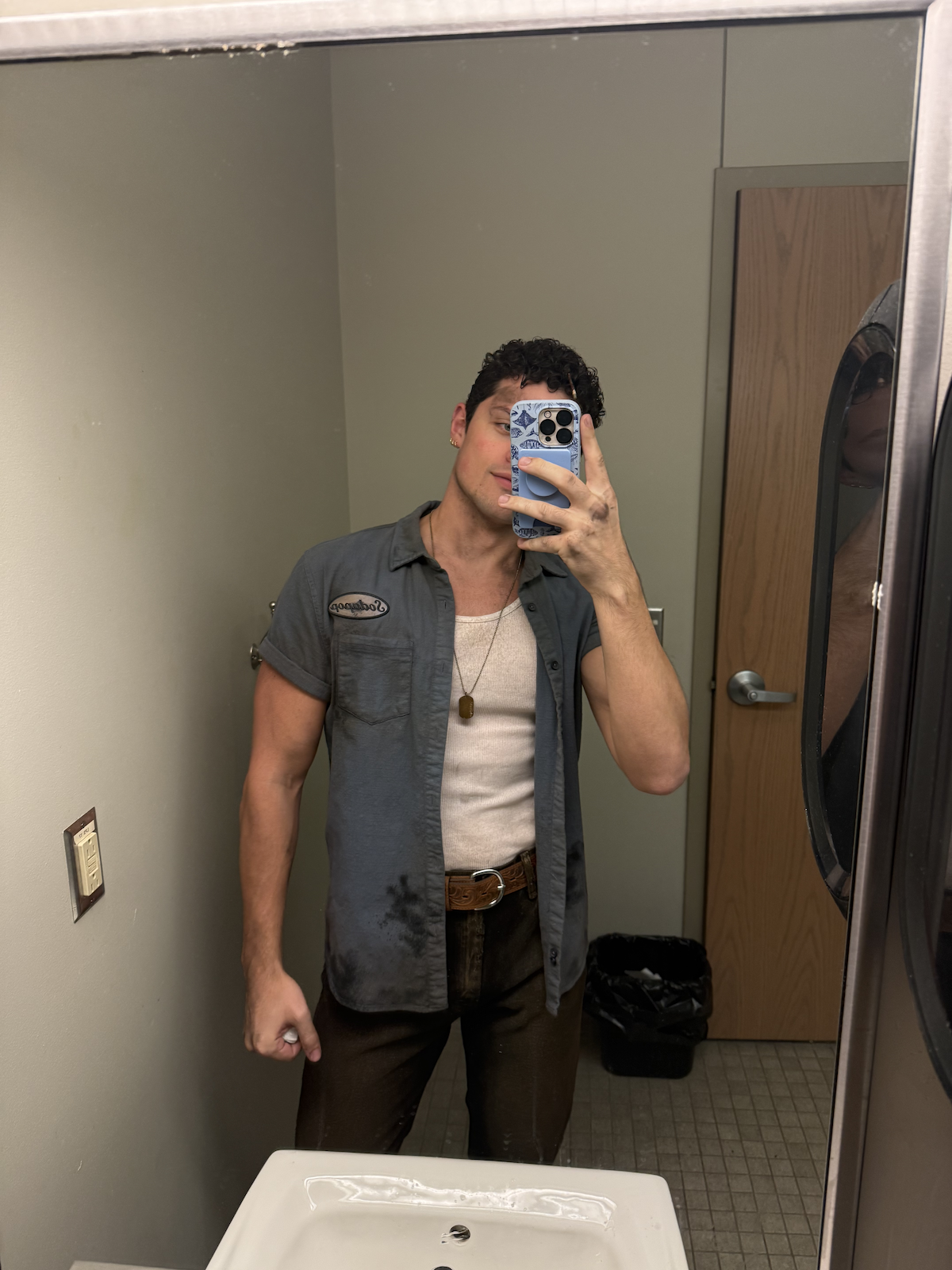 A young man taking a mirror selfie in a public restroom. He is wearing a gray short-sleeved shirt over a beige tank top, with dark pants and a belt. He has short curly hair and earrings, and is holding a phone with a blue and white patterned case. Th