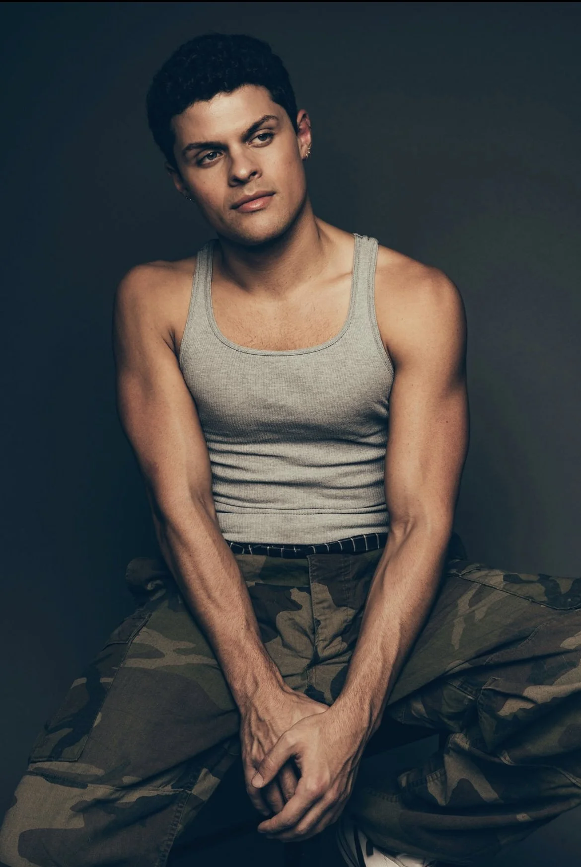 A young man with dark curly hair wearing a gray tank top and camouflage pants, sitting with his hands clasped and looking to the side against a dark background.