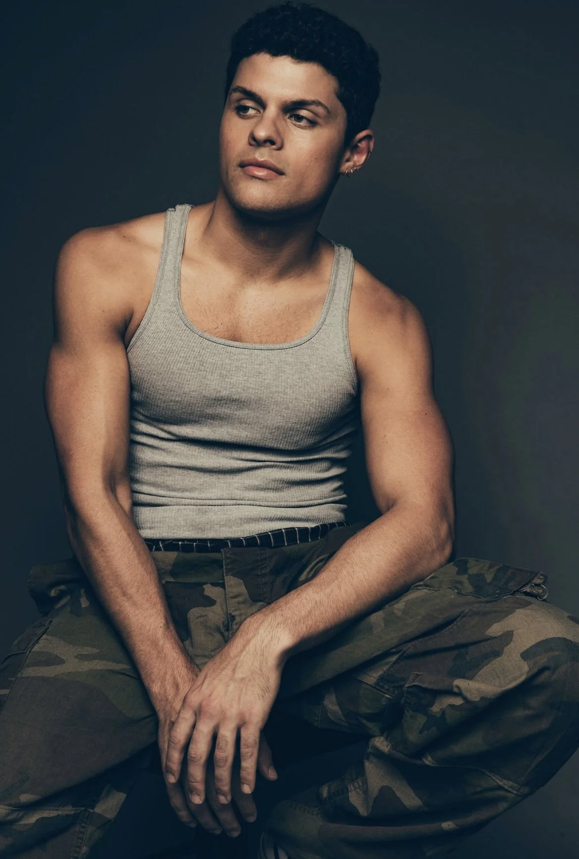 Young man with short dark curly hair wearing a gray sleeveless tank top and camouflage pants sitting against a dark background.