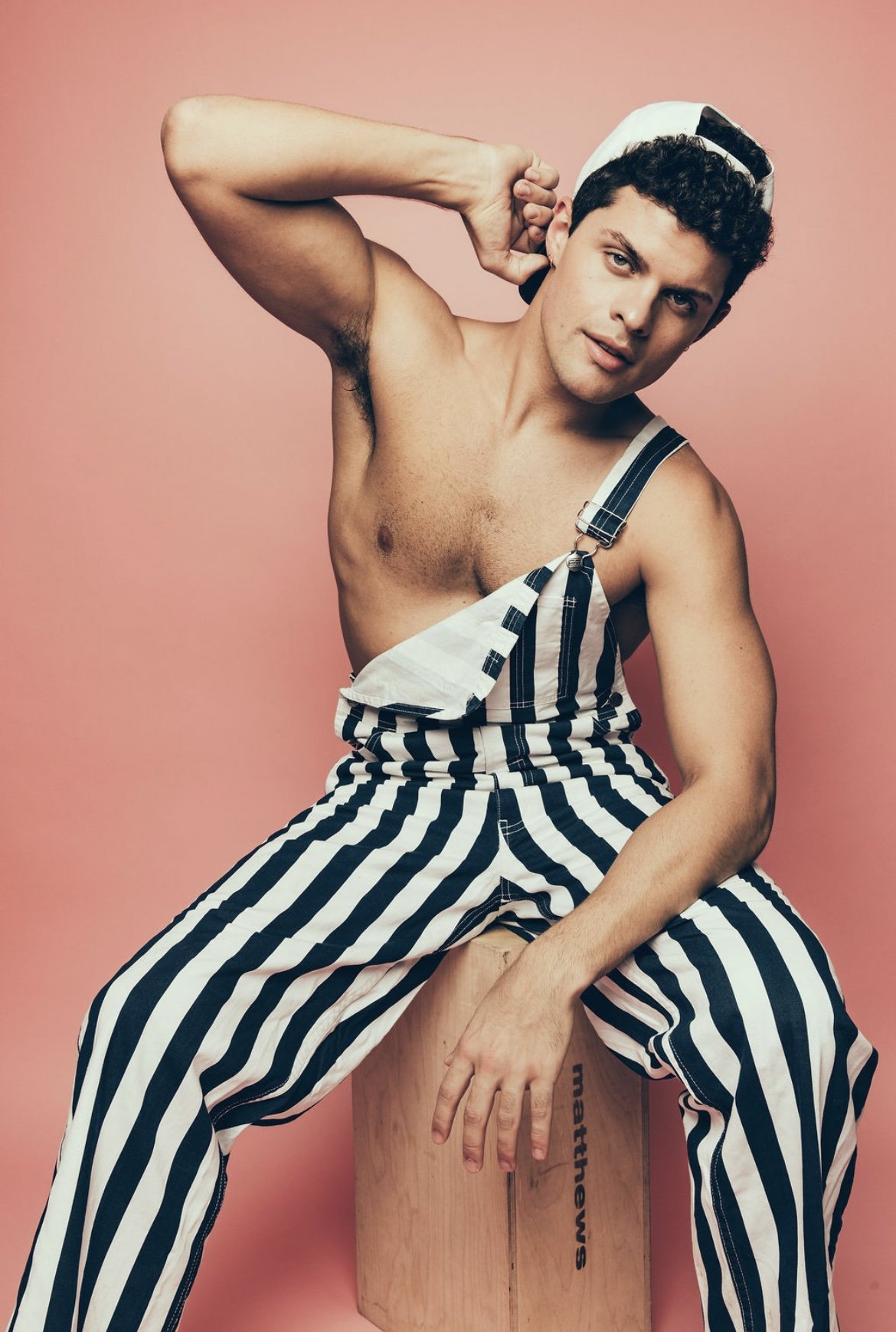 A shirtless young man wearing striped overalls and a backwards cap, sitting on a wooden box against a pink background, with one arm behind his head and one hand resting on his knee.
