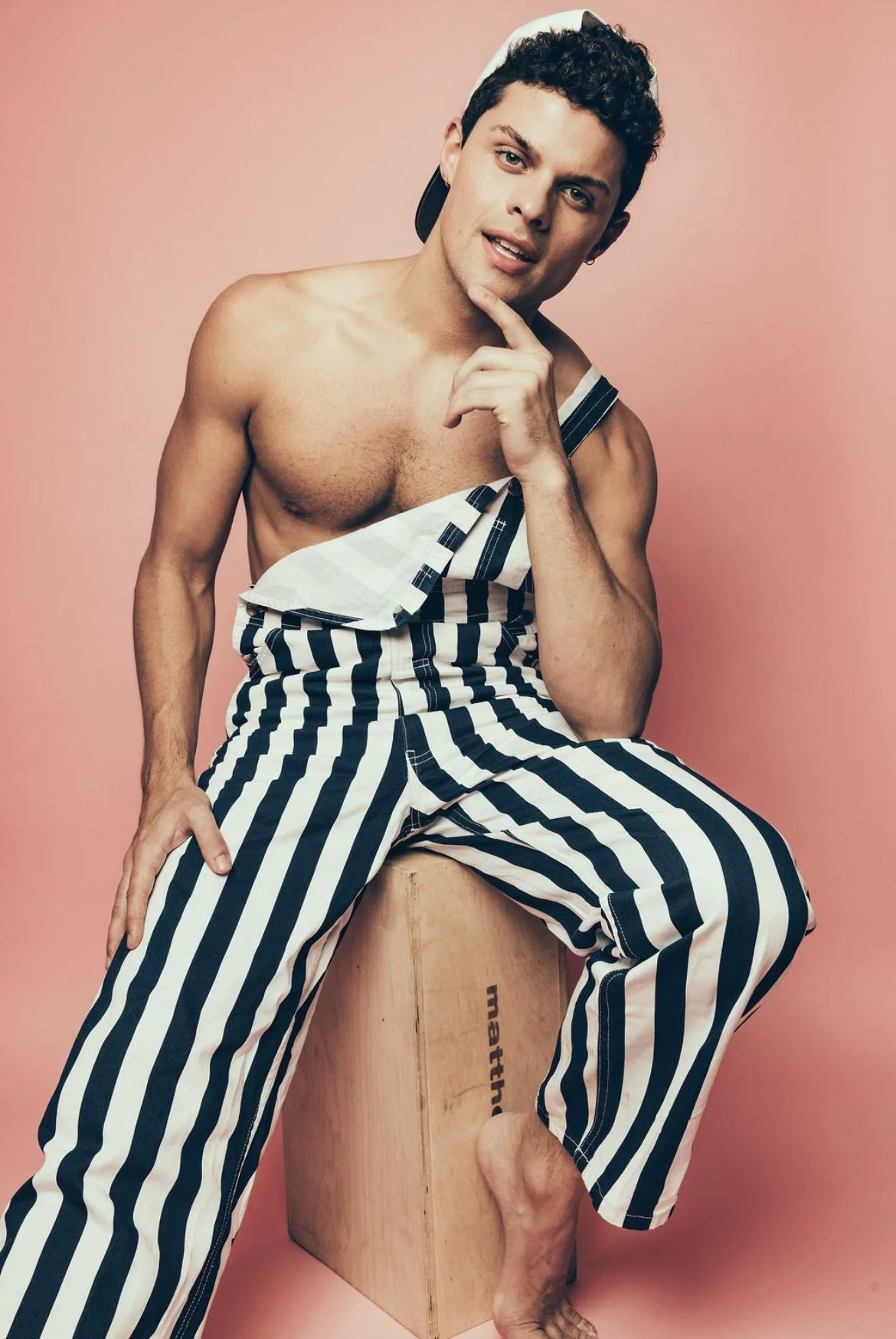 A young man with curly dark hair and light skin, wearing striped pants and a backwards cap, sitting barefoot on a wooden box with a pink background. He is shirtless, with one hand on his chin and the other on his thigh, looking at the camera with a s