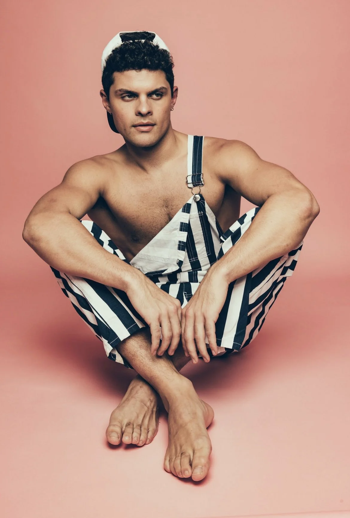 A young man with curly dark hair is sitting cross-legged on a pink background, wearing striped overalls pulled down to reveal his bare chest. He has a serious expression and is looking slightly to the side.
