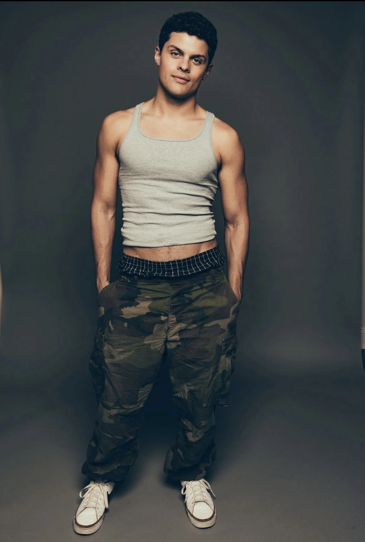Young man standing against dark background, wearing a gray tank top, camouflage pants, and white sneakers.