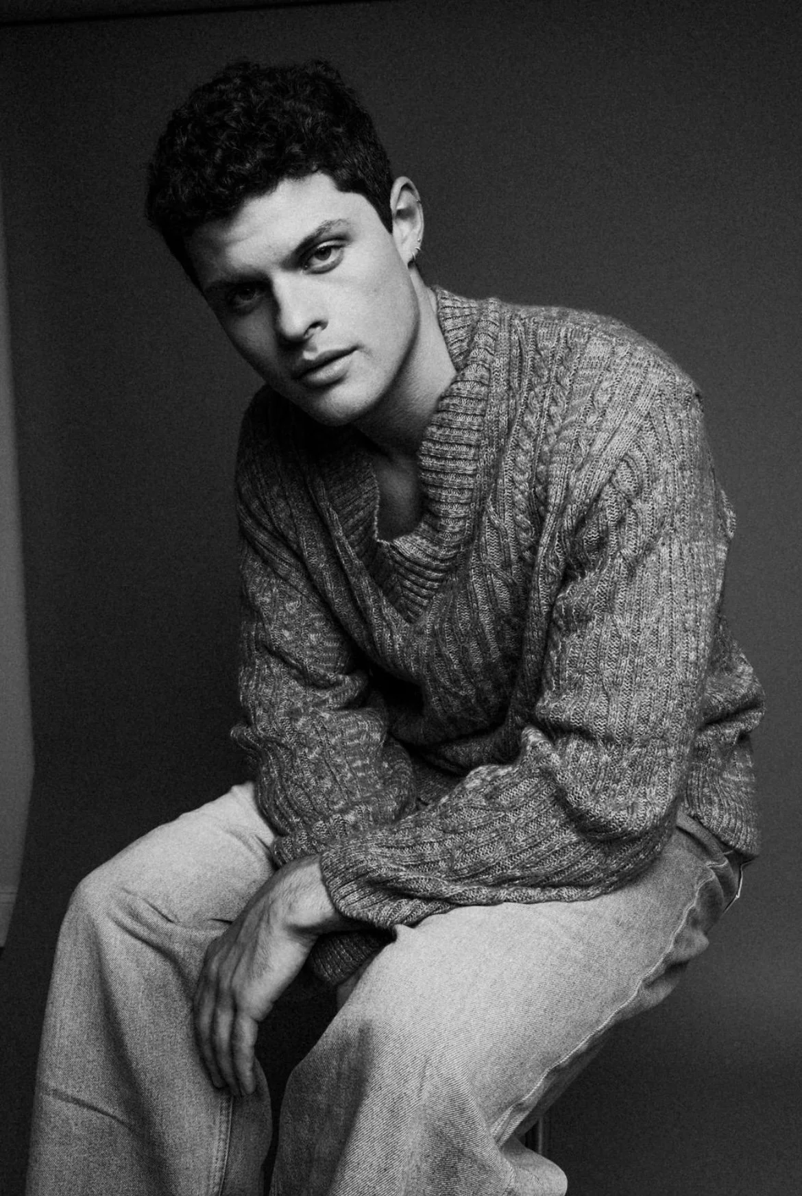 Black and white photo of a young man with short curly hair, wearing a textured knit sweater and light-colored pants, sitting with a relaxed posture against a plain dark background.