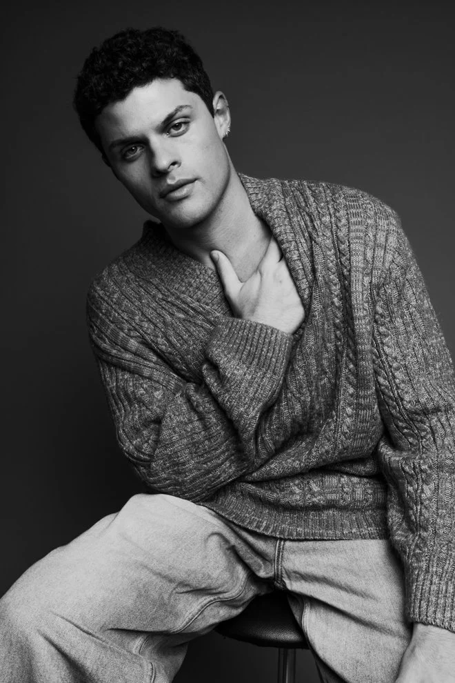 A young man with curly hair, wearing a textured sweater and light-colored pants, sitting on a stool against a plain background, looking at the camera in a black and white portrait.