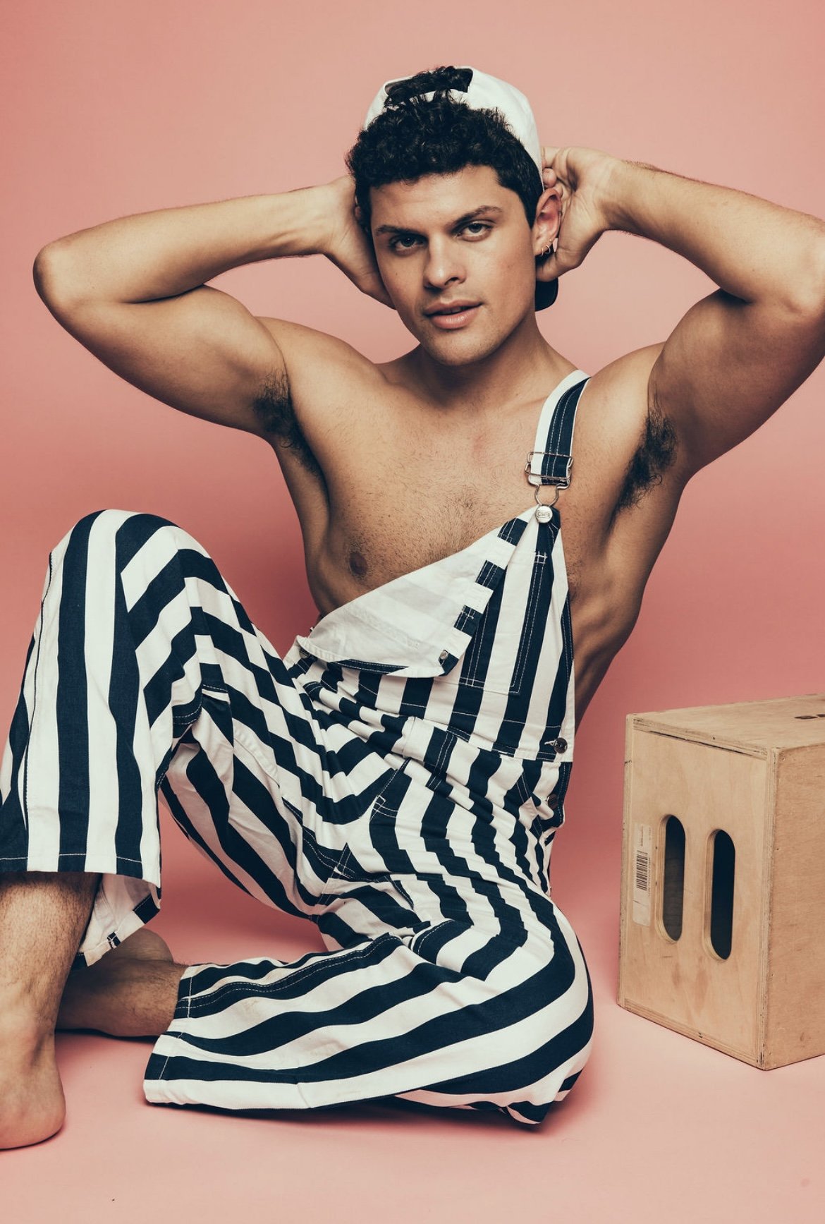 A young man with curly hair is shirtless and wearing striped overalls. He is posing with his hands behind his head, looking directly at the camera against a pink background. There is a wooden crate on the floor next to him.