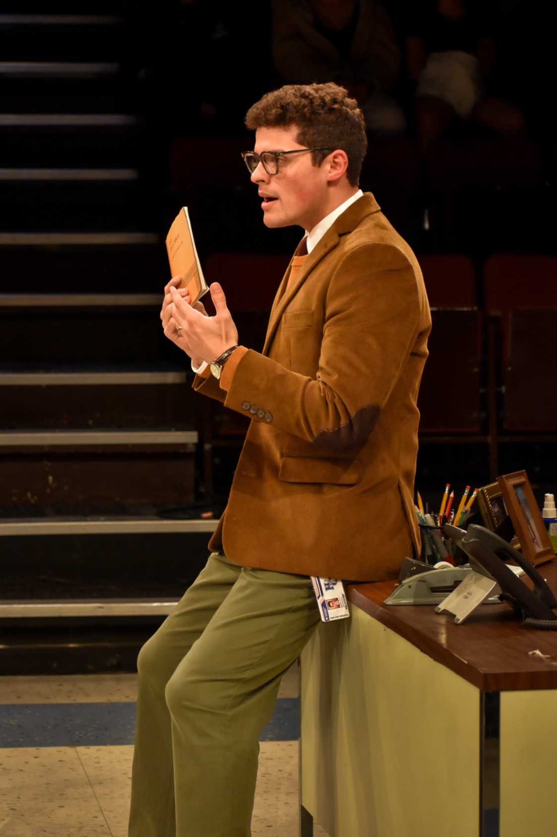 A man wearing glasses, a brown blazer, white shirt, and khaki pants is standing and reading a book in a classroom or lecture hall. There are desks, supplies, and a few people seated in the background.