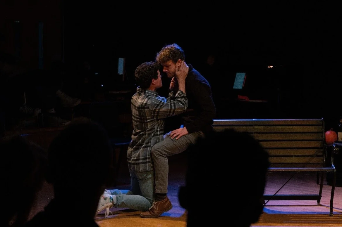 Two men sharing an intimate moment on stage, with one kneeling and the other sitting on a bench, in a dimly lit theater or performance space.