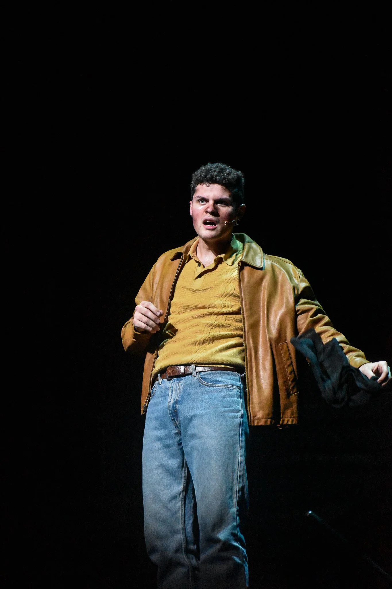 A young man on stage, wearing a brown leather jacket, yellow shirt, and blue jeans, appears to be speaking or performing, with a dark background.