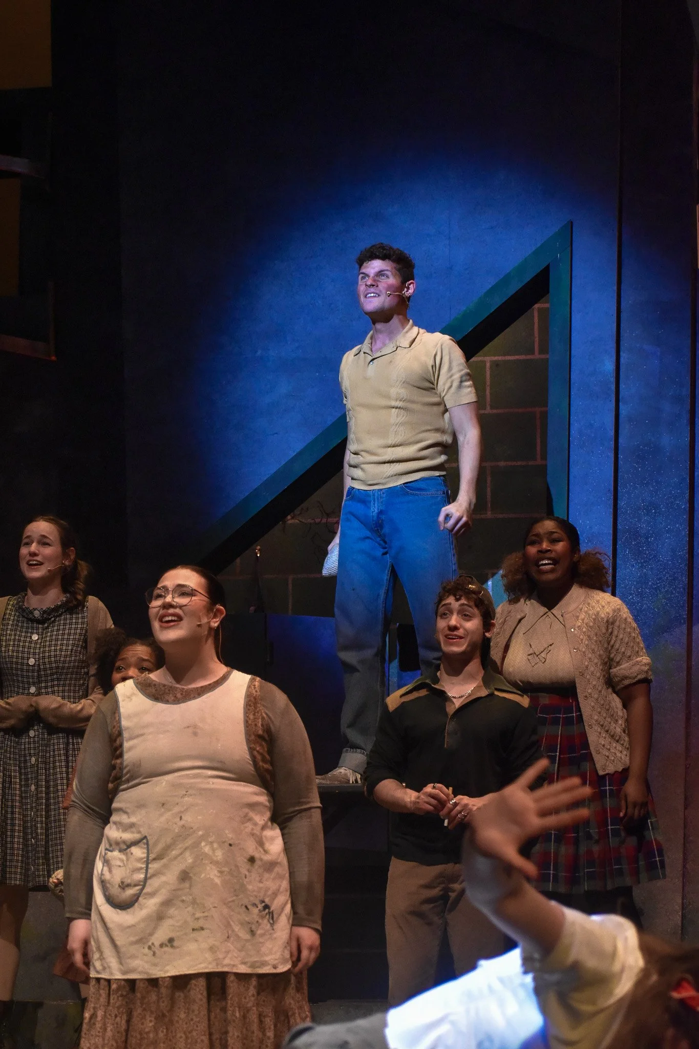 A group of people on stage, with one young man in the center wearing a yellowish-brown shirt and blue jeans, smiling and looking upward. Others around him are also smiling and looking in various directions. The background has a blue wall with stairs 