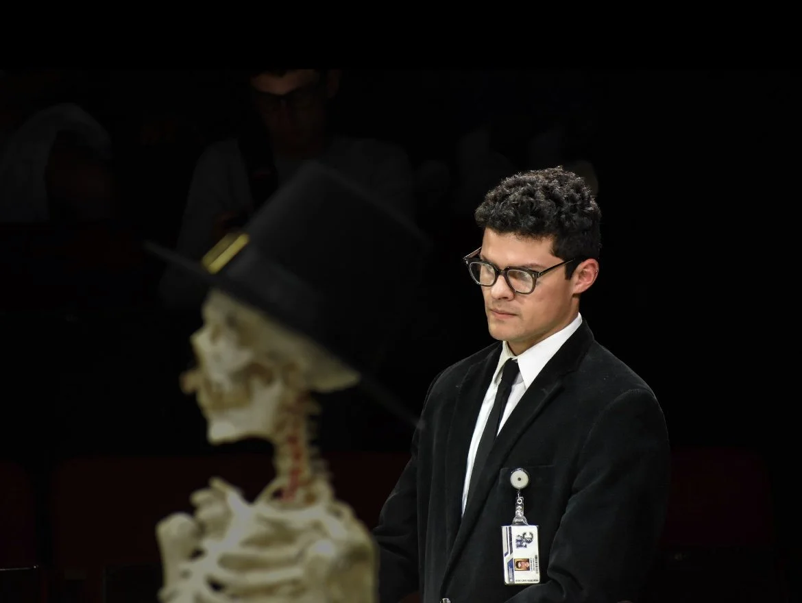 A man wearing glasses, a black velvet jacket, white shirt, and black tie stands with his eyes closed as a skeleton in a top hat looks at him, on a dark background.