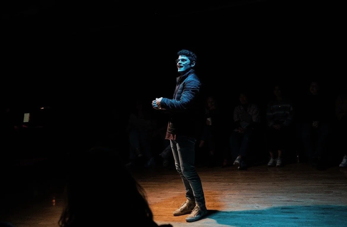 A young man standing on a stage in a spotlight during a performance, with a dark background and an audience in the shadows.