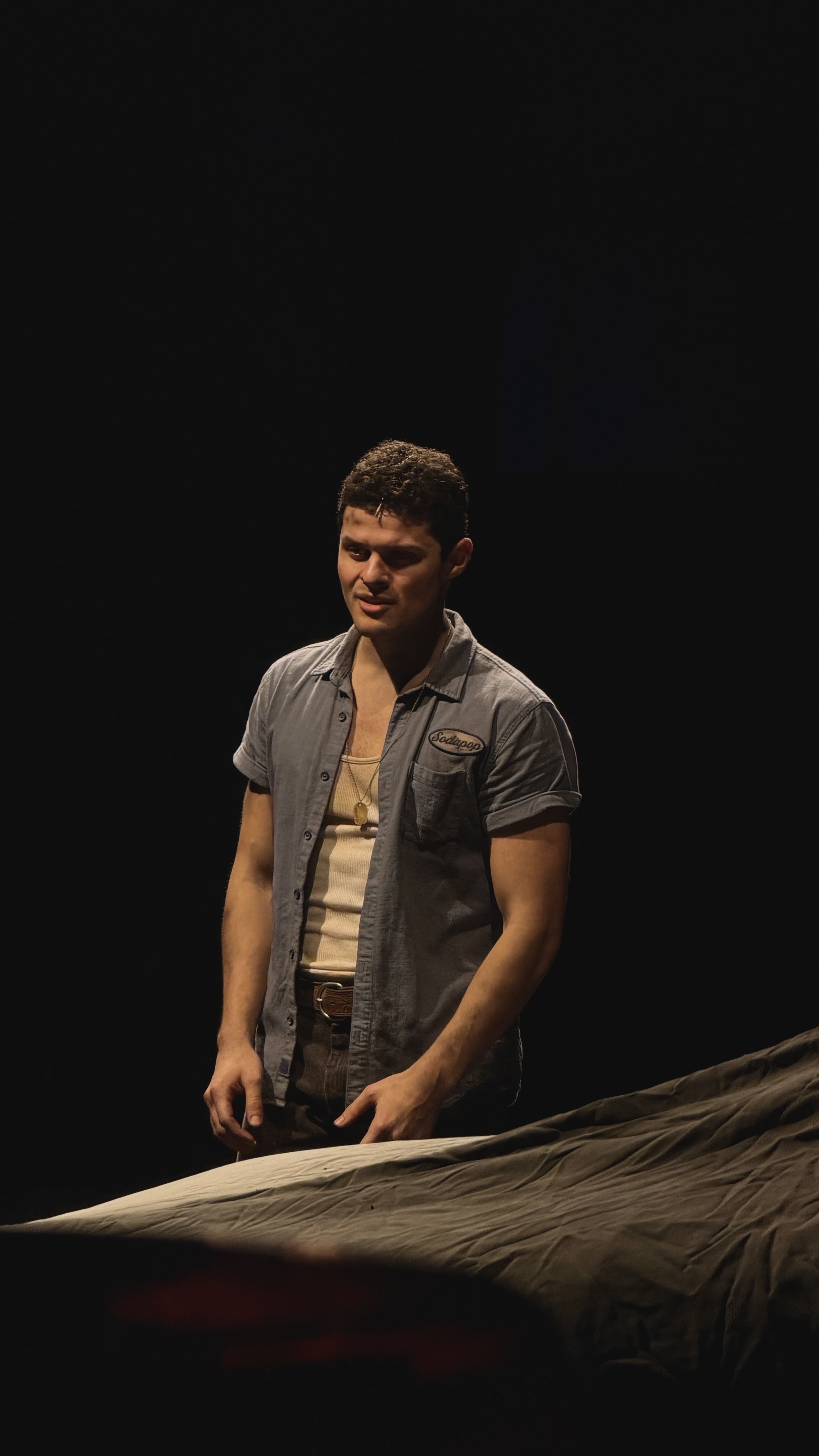 A young man with curly dark hair stands in a mostly dark room with a single light illuminating his face and upper body. He is wearing a grey short-sleeved shirt over a beige tank top, with a necklace. He is looking slightly downward, with a slight sm