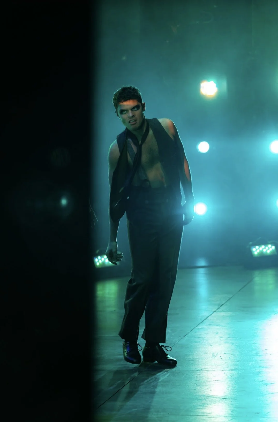 A man performing on stage with dramatic lighting, shirtless and wearing dark pants, some of his clothing is hanging off his shoulders, and he has a serious expression.