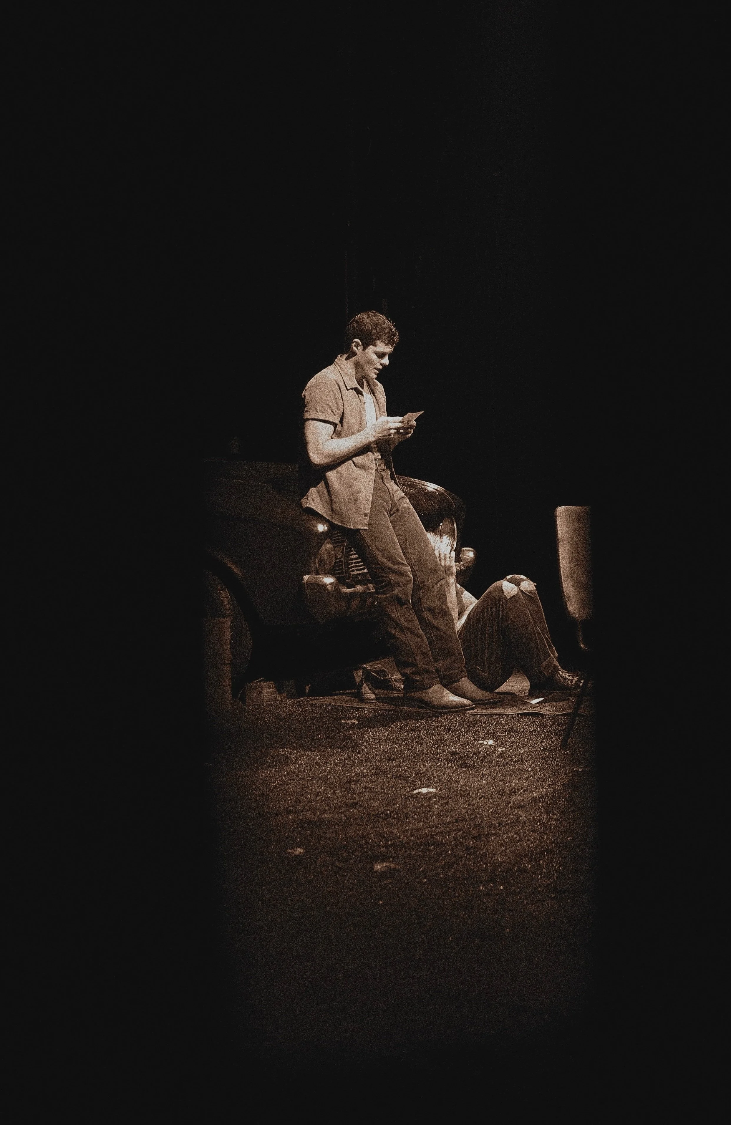 A young man sitting on a car at night, looking at his phone, with another person lying on the ground nearby.