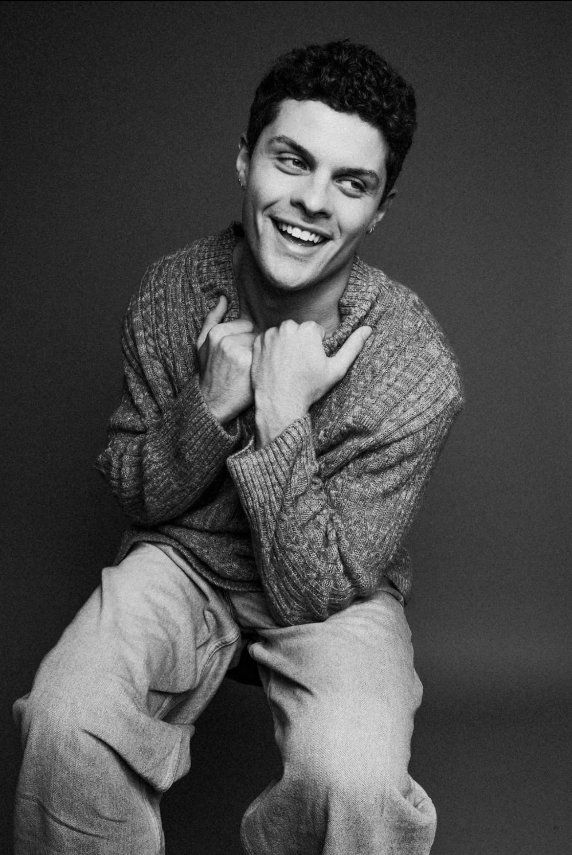Black and white photo of a young man sitting and smiling with his hands crossed over his chest, wearing a knit sweater and light-colored pants, against a plain dark background.