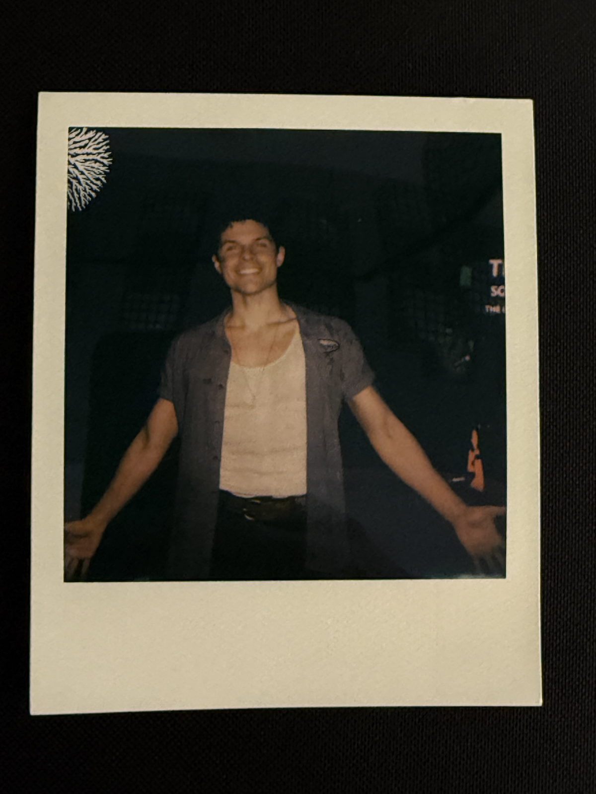 A young man with dark hair smiling with his arms outstretched is standing in a dark setting, wearing a white t-shirt, a grey unbuttoned shirt, and a necklace.