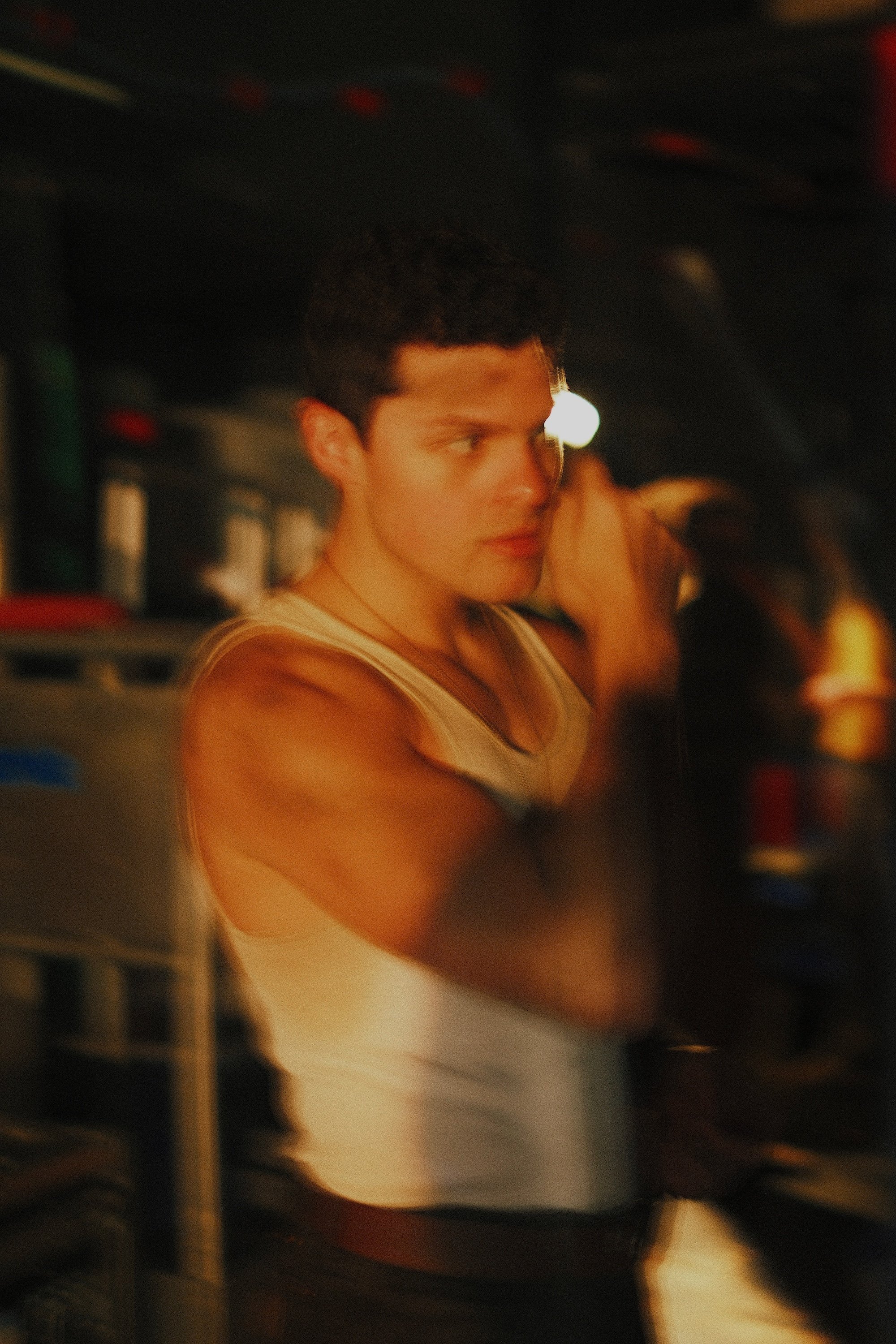 A young man with dark, curly hair and light skin is in a dimly lit environment, looking to his right. He is wearing a cream-colored tank top and appears to be adjusting his hair or left ear. The background is dark with some lights and objects slightly visible.