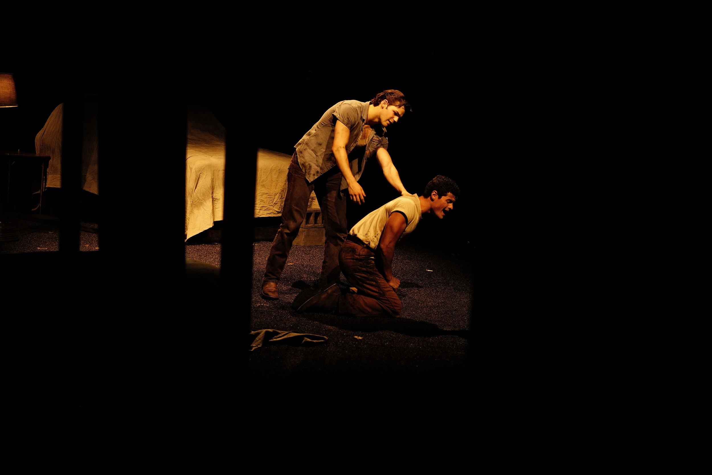 Two men are on stage, one standing and one kneeling, in a theatrical performance with a dark background, a bed, and a nightstand visible.
