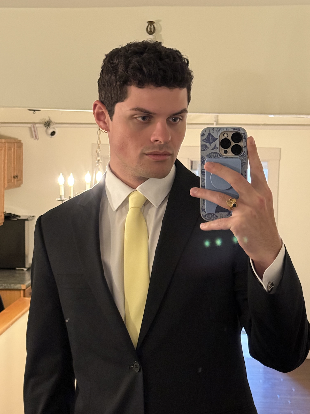 A man taking a mirror selfie, dressed in a black suit with a white shirt and yellow tie, in a room with warm lighting and a chandelier, holding a smartphone with a patterned case.