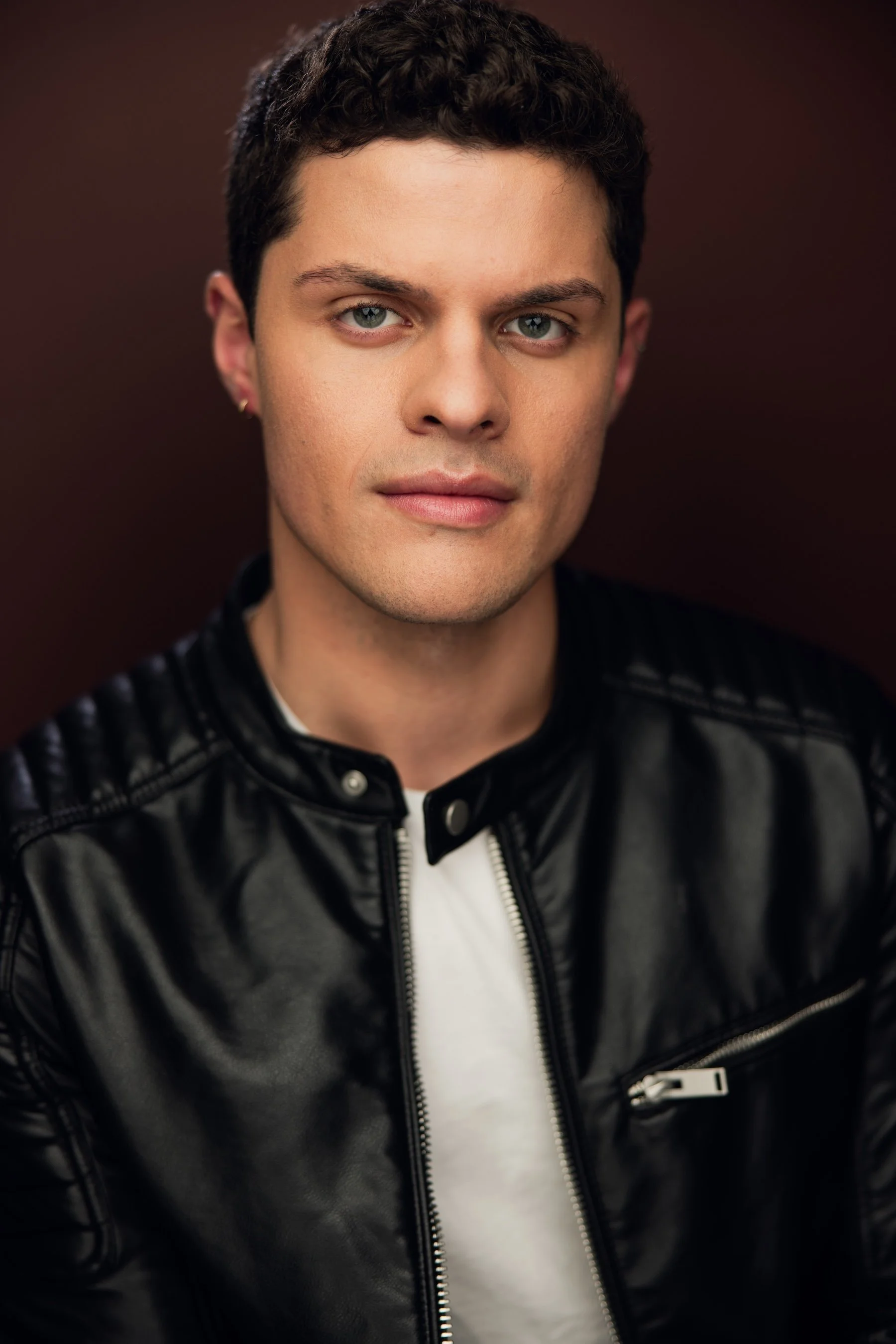 Close-up portrait of a young man with short dark curly hair and blue eyes, wearing a black leather jacket over a white shirt, against a dark background.