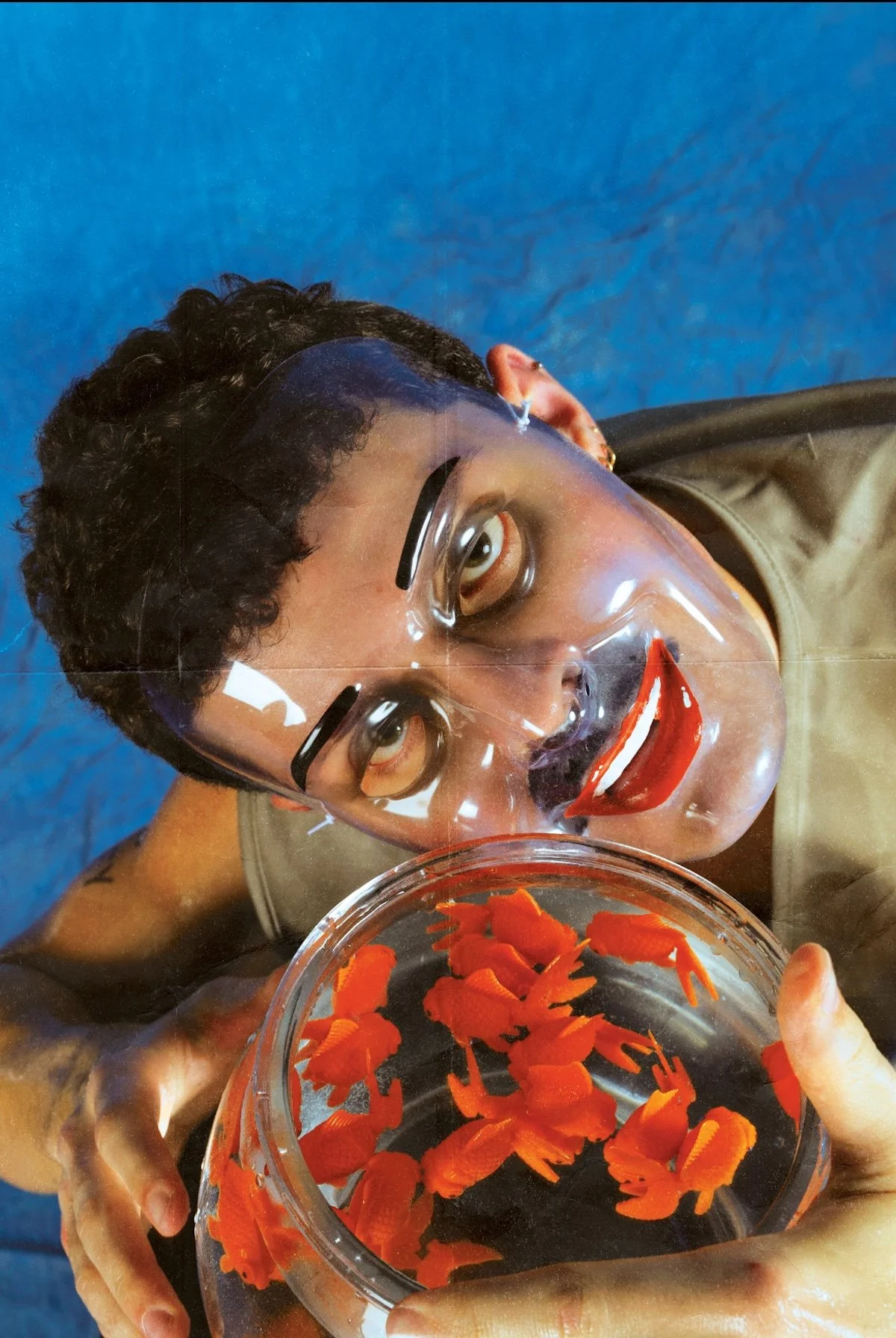 A person with short curly hair wearing a clear face shield and glasses, smiling with red lipstick, holding a glass bowl of orange flower petals, with a blue background.