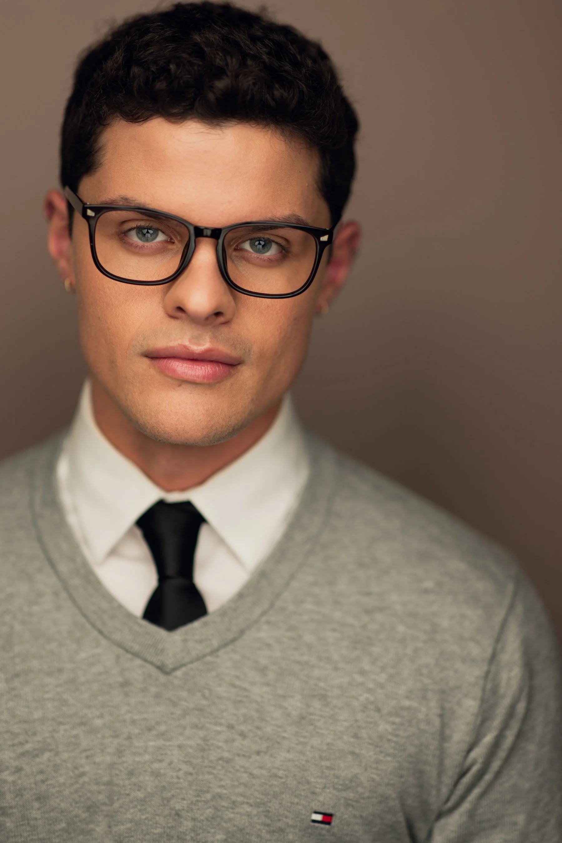 Close-up of a young man with short dark curly hair, wearing black-rimmed glasses, a white collared shirt, a gray sweater with a small Tommy Hilfiger logo, and a black tie, against a plain background.