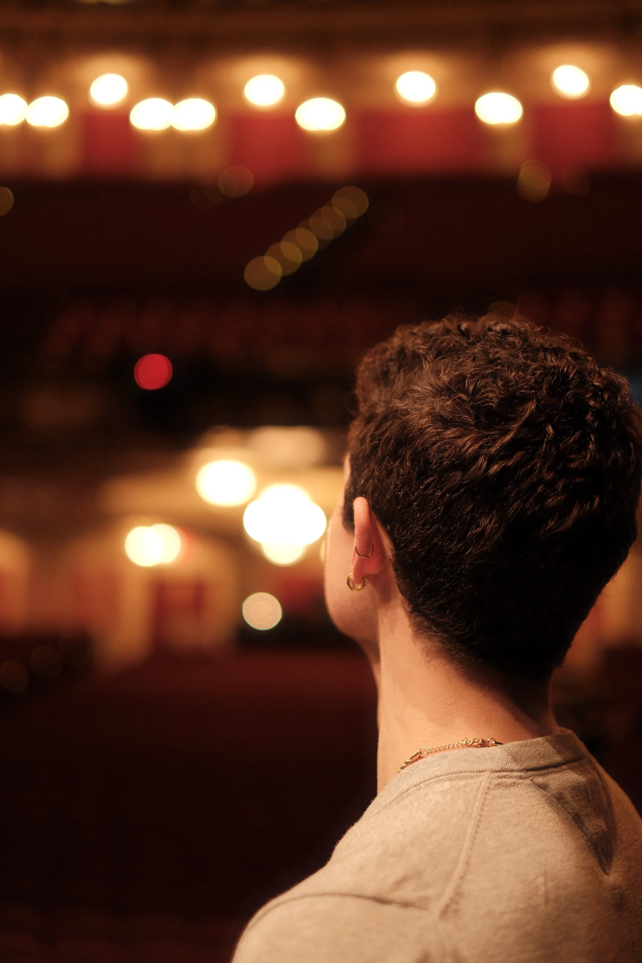 Back view of a person with short, curly hair, earrings, and earrings, wearing a beige shirt, with bokeh lights in the background.