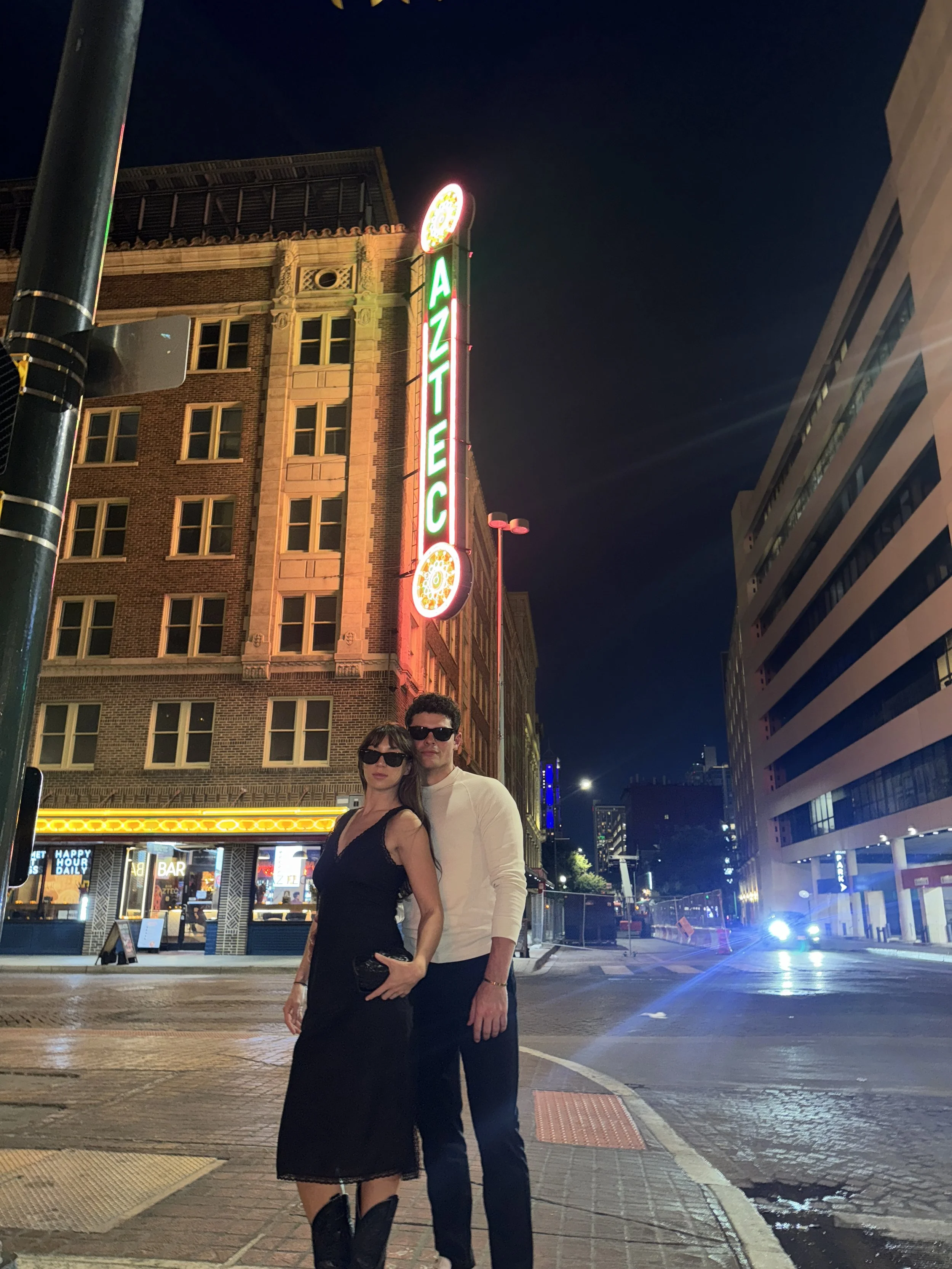 A man and woman standing on an illuminated city street at night, with neon signs and tall buildings in the background. The woman is wearing a black dress and knee-high boots, and the man is wearing sunglasses and a white shirt.
