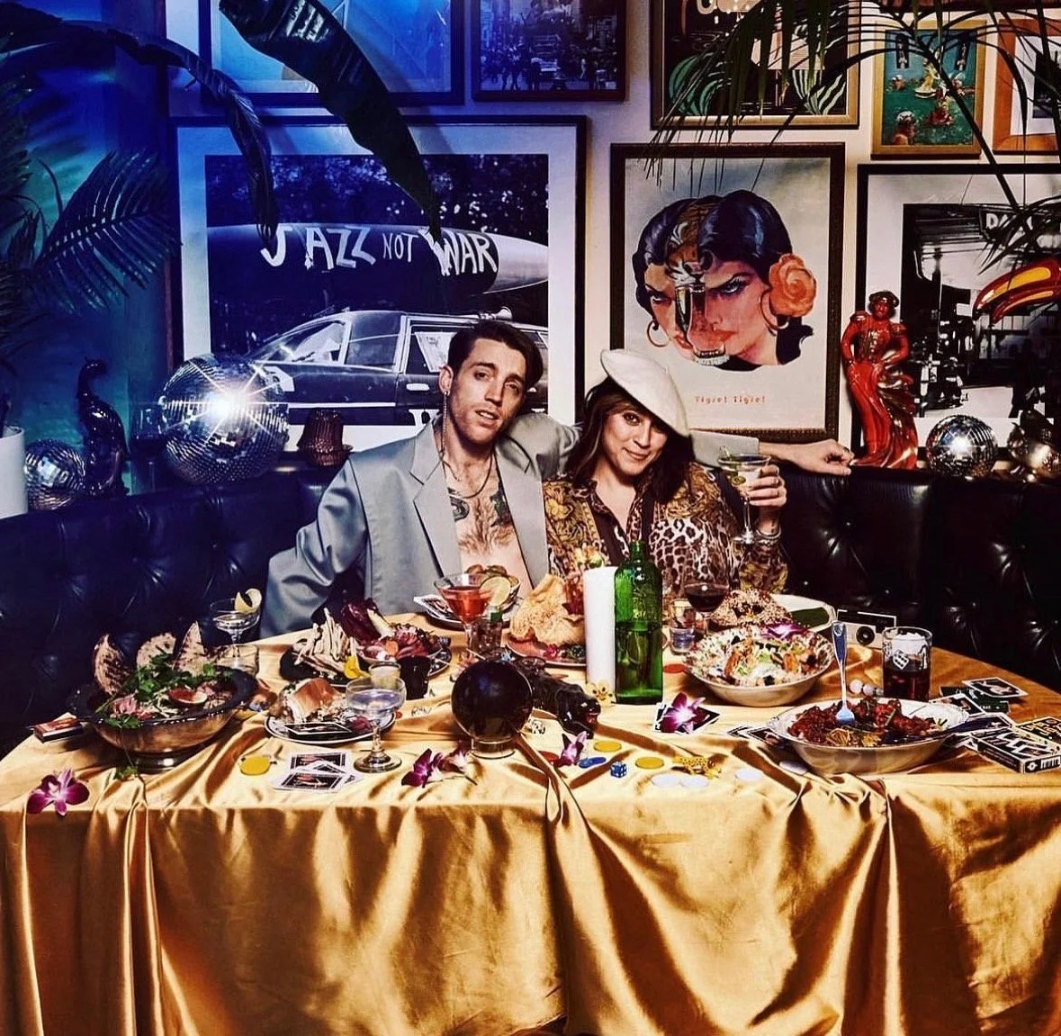 Two people sitting at a decorated dinner table in a colorful, art-filled room. The man is wearing a shiny gray jacket, and the woman is wearing a leopard print outfit with a white hat. The table has various dishes, drinks, and decorative items, with posters and artwork on the wall behind them.
